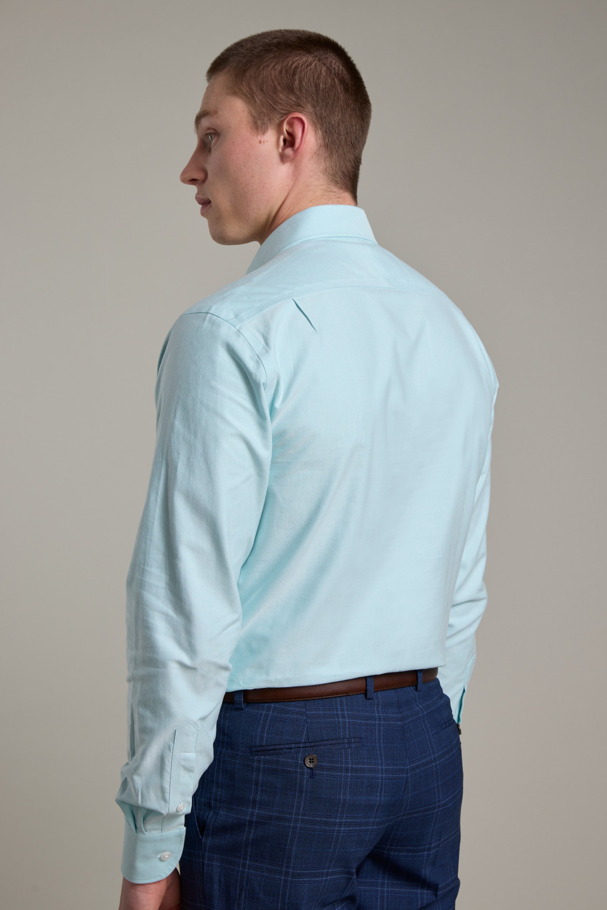 A man with short brown hair, wearing the Barkers Edgar Texture Business Shirt in light blue and blue plaid pants, stands facing away from the camera against a plain, light background.