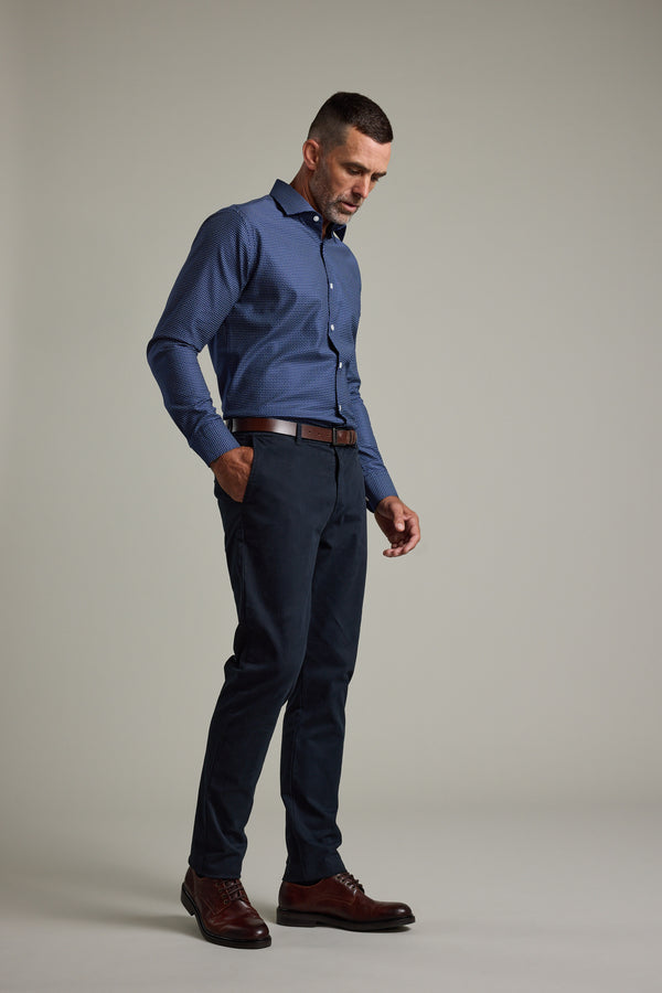 A man stands against a plain background wearing the Barkers Maiden Paisley Business Shirt, dark pants, a brown belt, and brown dress shoes. He looks down with one hand in his pocket.