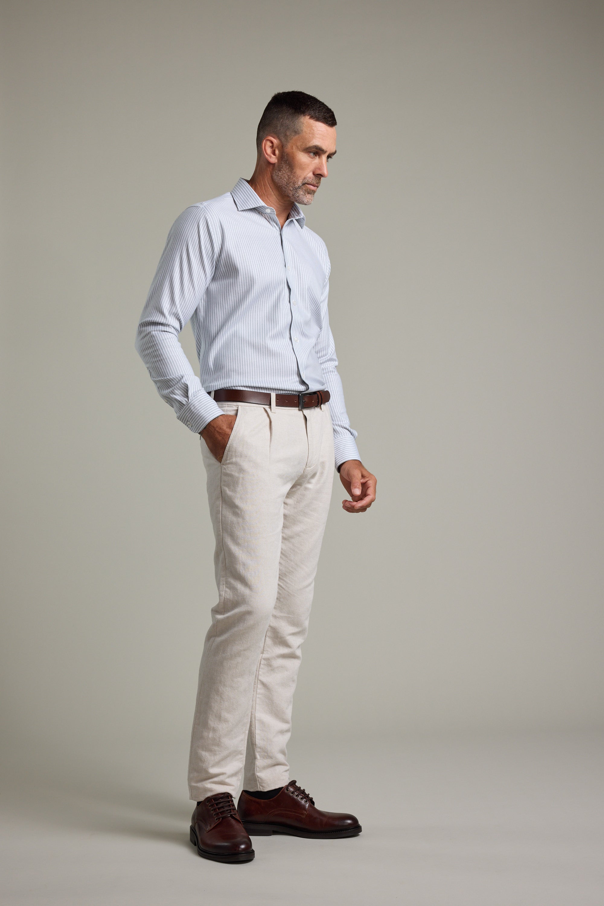 A man with short hair and a beard stands against a plain background wearing the Barkers Clarke Stripe Business Shirt, beige trousers, a brown belt, and dark brown dress shoes. He has one hand in his pocket and a thoughtful expression.