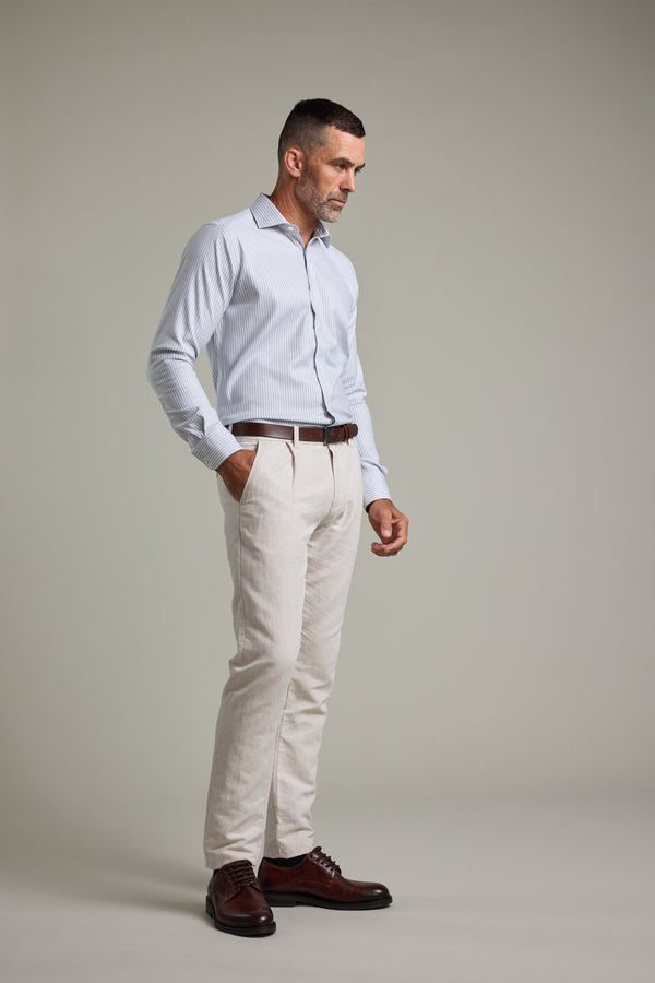 A man with short hair and a beard stands against a plain background wearing the Barkers Clarke Stripe Business Shirt, beige trousers, a brown belt, and dark brown dress shoes. He has one hand in his pocket and a thoughtful expression.