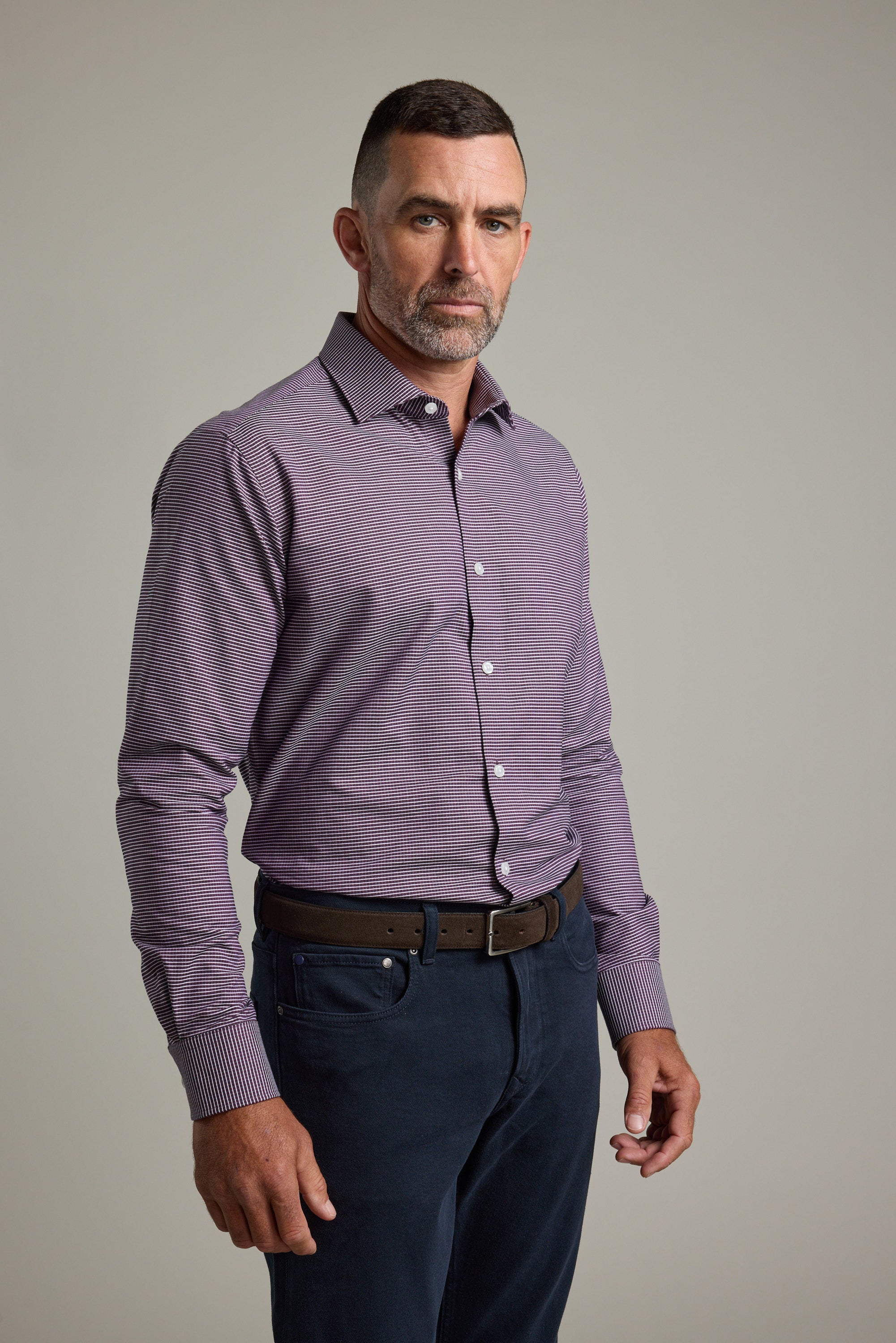 A man with short dark hair and a trimmed beard stands against a plain background, wearing the Barkers Waverly Check Business Shirt, dark pants, and a brown belt. He looks directly at the camera with a neutral expression.