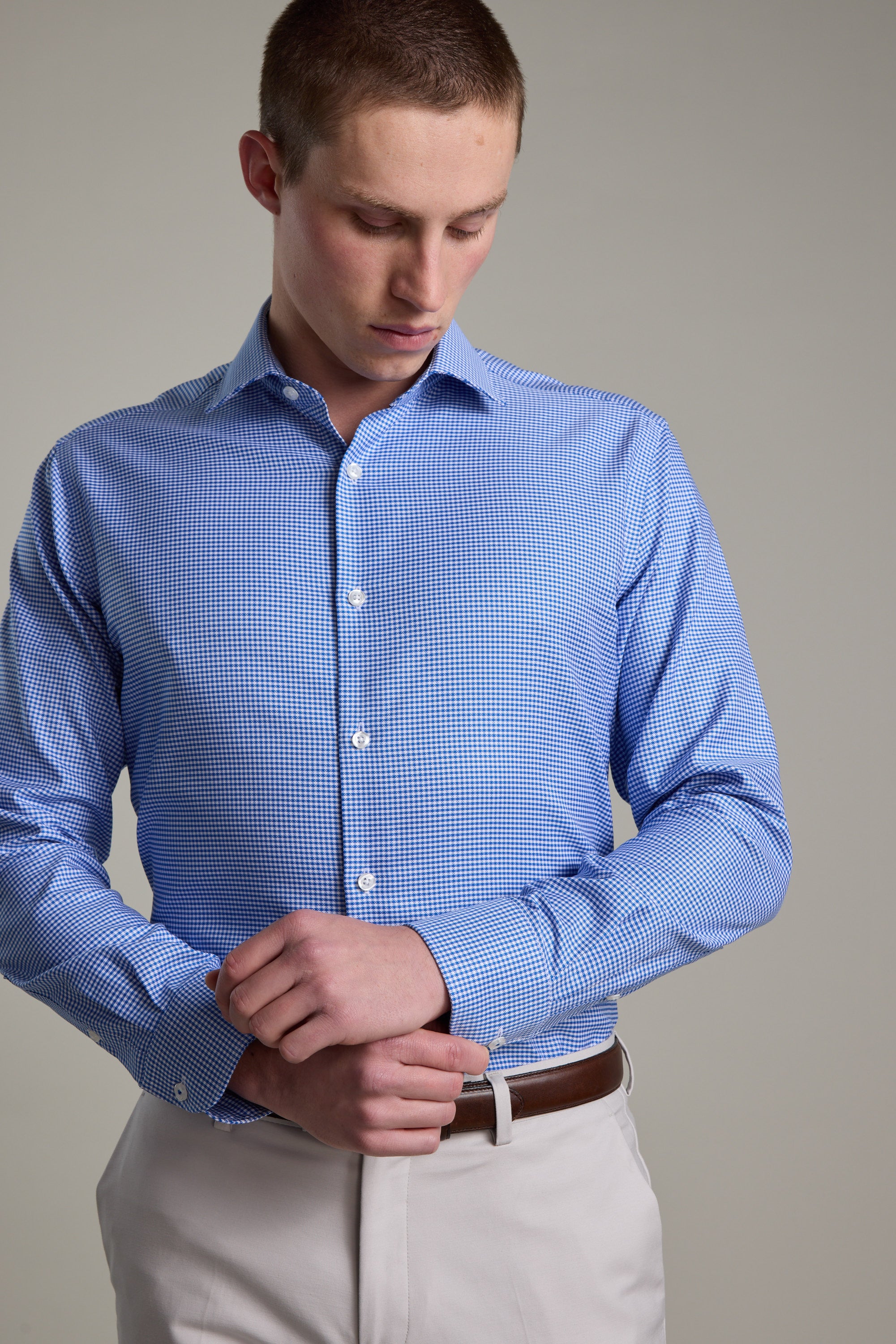 A man in a Barkers Mitchel Check Business Shirt—a light blue, tailored fit with a checkered pattern—and beige pants with a brown belt looks down as he adjusts his left shirt cuff against a plain background.