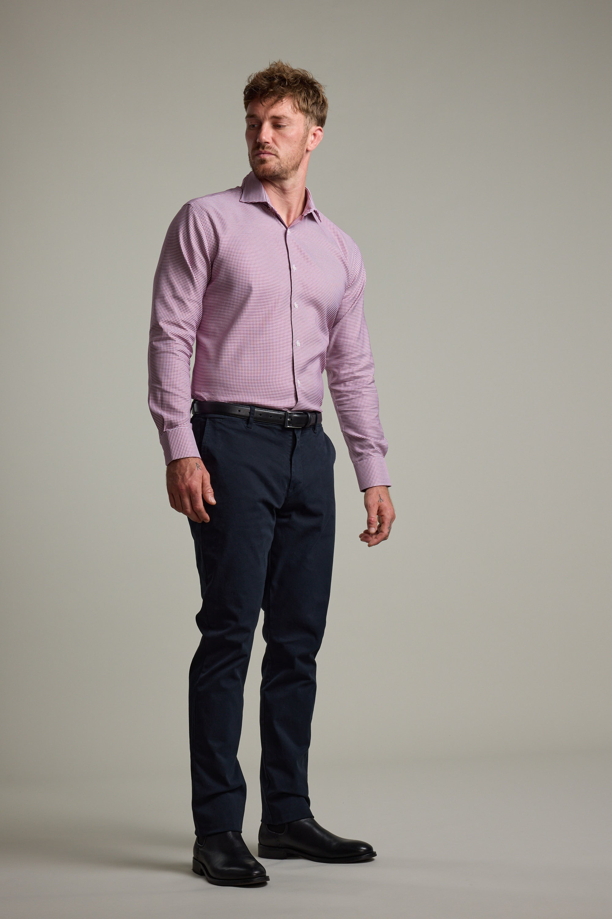 A man stands slightly turned, wearing the Barkers Mitchel Check Business Shirt with dark trousers and black shoes against a plain, light background.