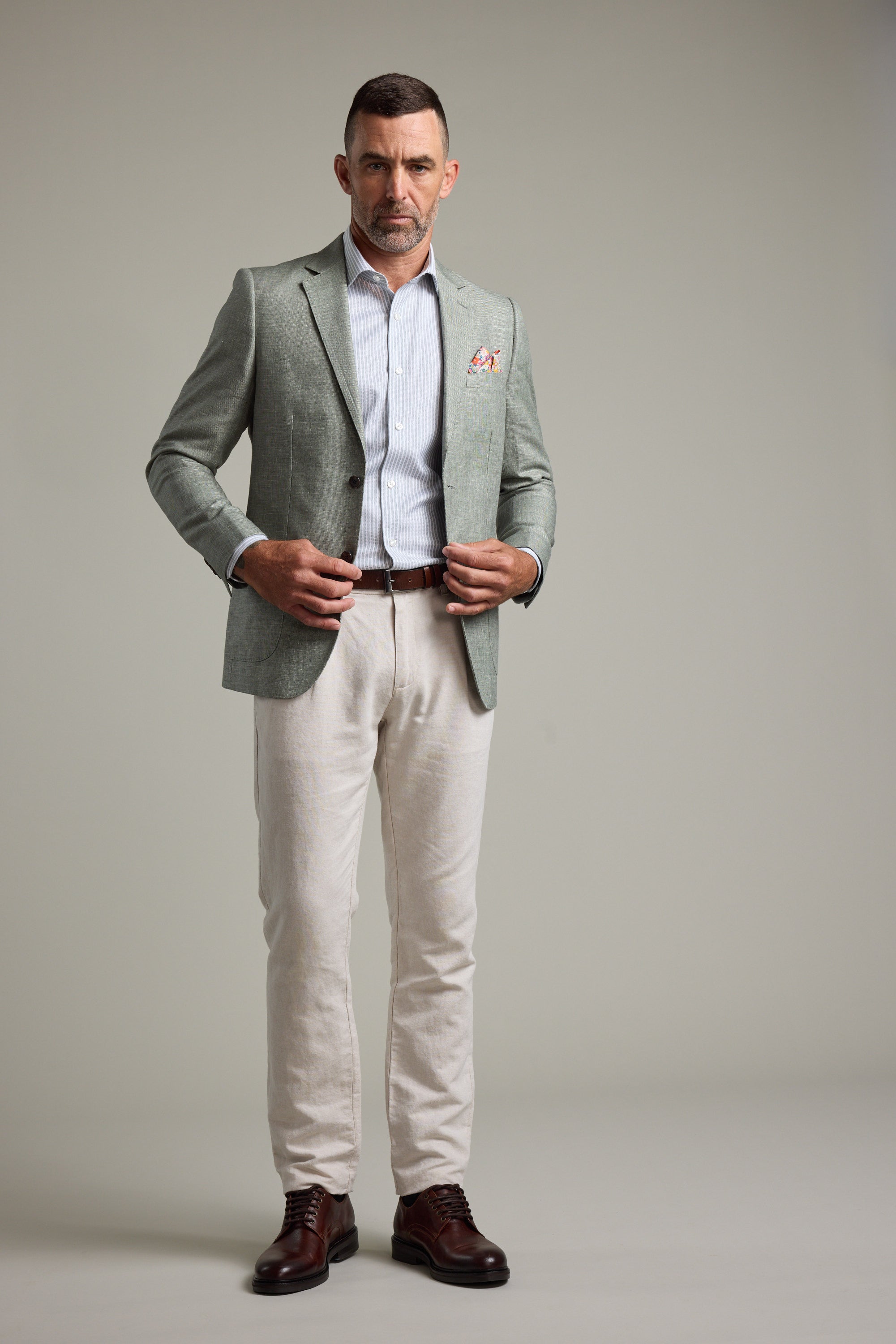 A man stands facing forward in a Barkers Marion Texture Blazer, striped dress shirt, light beige trousers, and dark brown dress shoes against a neutral grey background—a perfect summer occasion outfit.