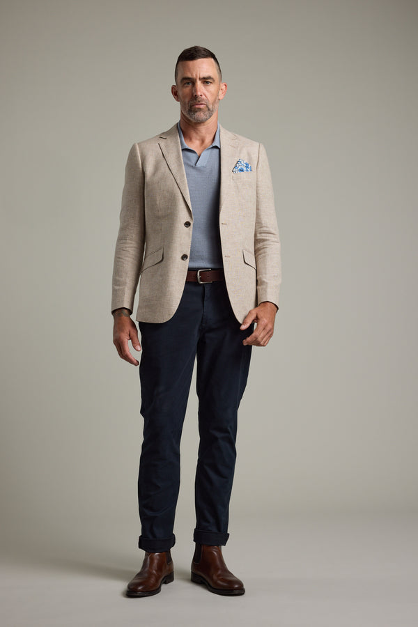 A man with short hair and a trimmed beard stands against a plain background, wearing Barkers' Welland Texture Blazer in beige with a blue pocket square, paired with a light blue polo shirt, dark blue pants, brown belt, and brown boots.