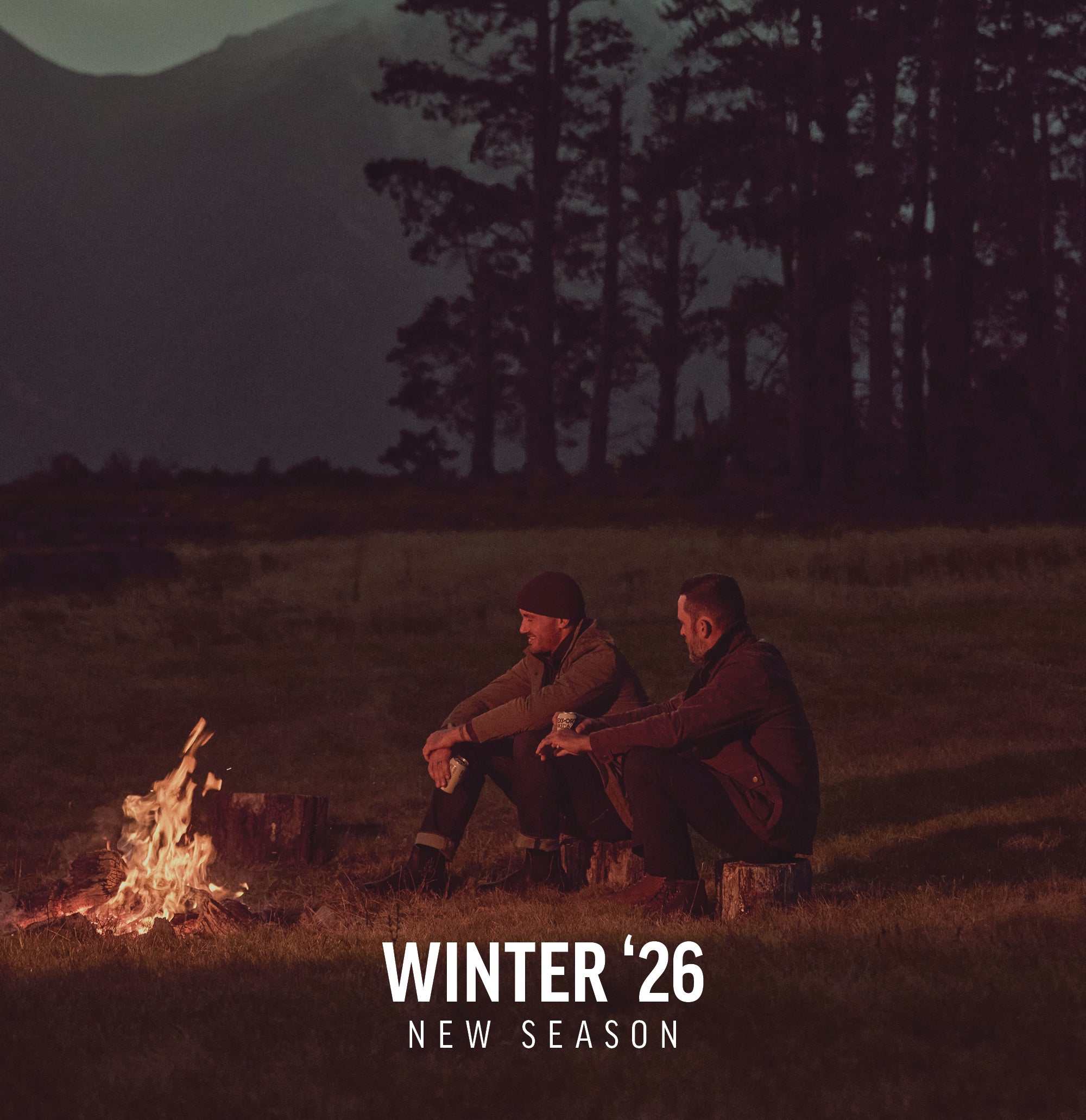 Two people sit by a campfire at dusk with NZ mountains and trees in the background.
