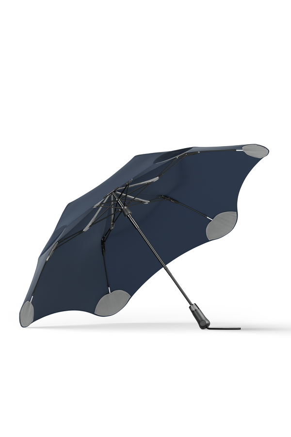 The Blunt Metro Umbrella by Blunt is shown open and angled to the side against a white background, featuring a dark blue color, aerodynamic design with rounded edges, and reinforced gray tips.