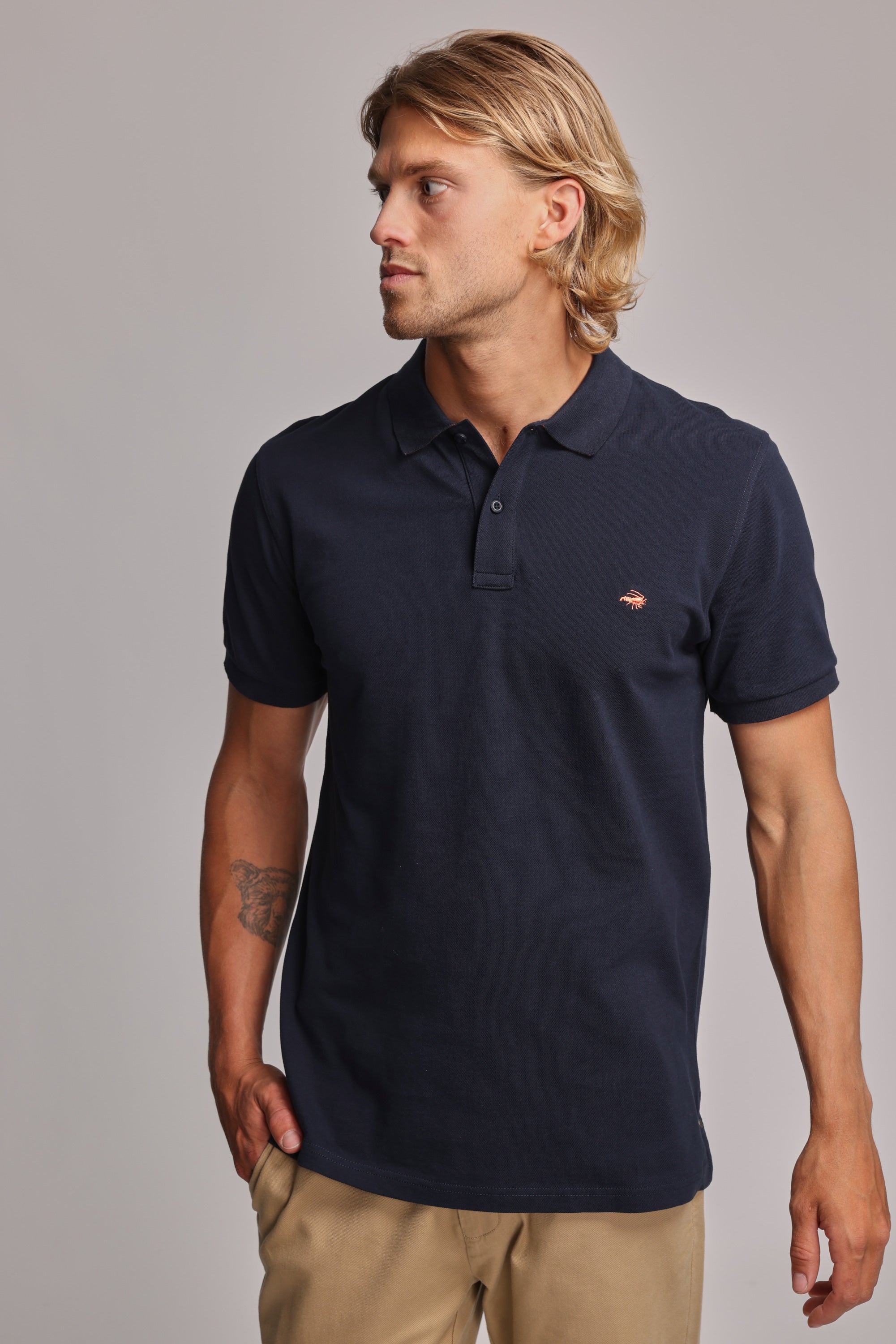 A man with shoulder-length blonde hair wears a navy Barkers Regent Pique Polo and khaki pants, standing against a plain light gray background and looking to his left with one hand in his pocket.