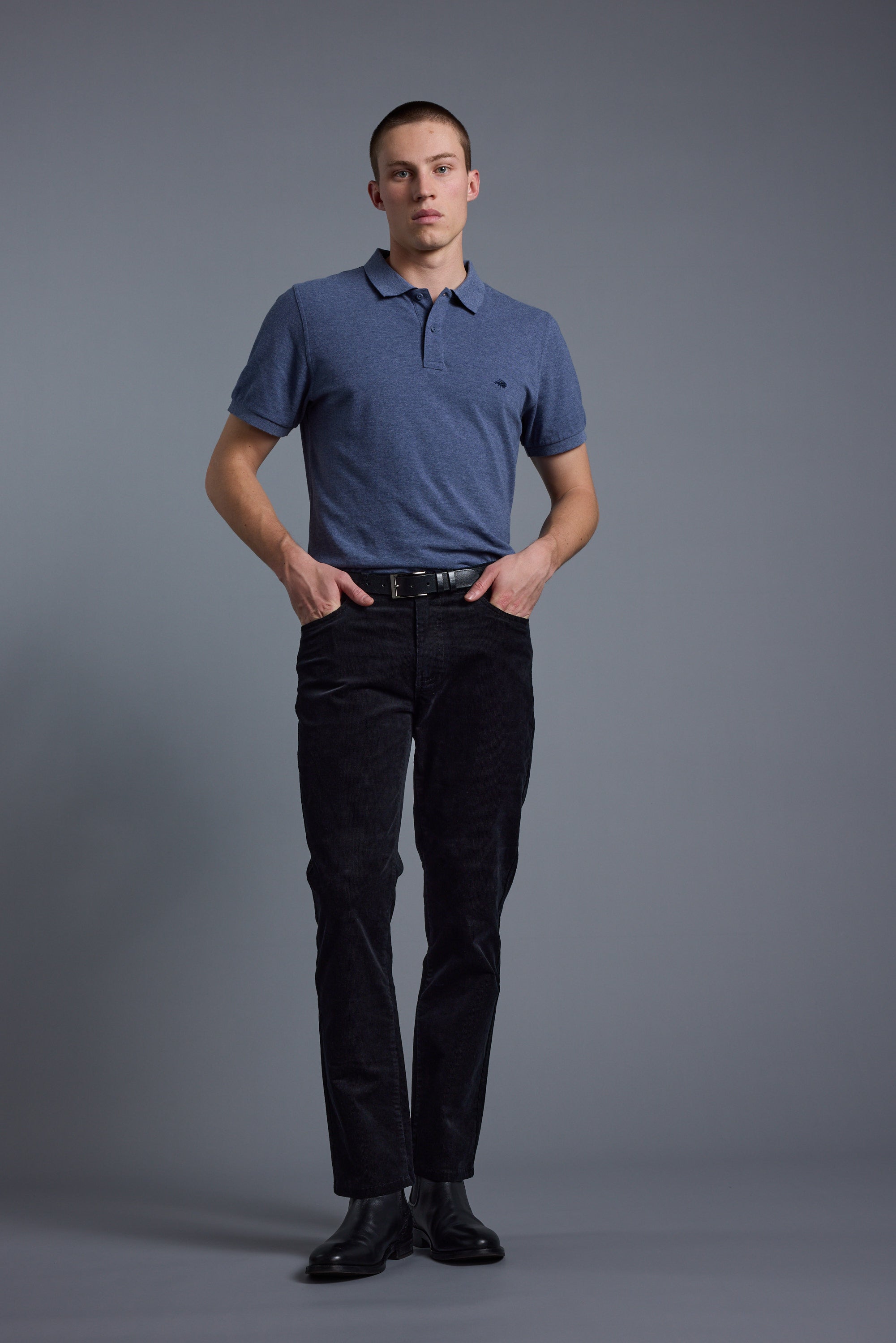 A young man stands against a plain gray background, wearing a Barkers Regent Pique Polo in blue, paired with black pants, a black belt, and black shoes. He has his hands in his pockets and maintains a neutral expression.