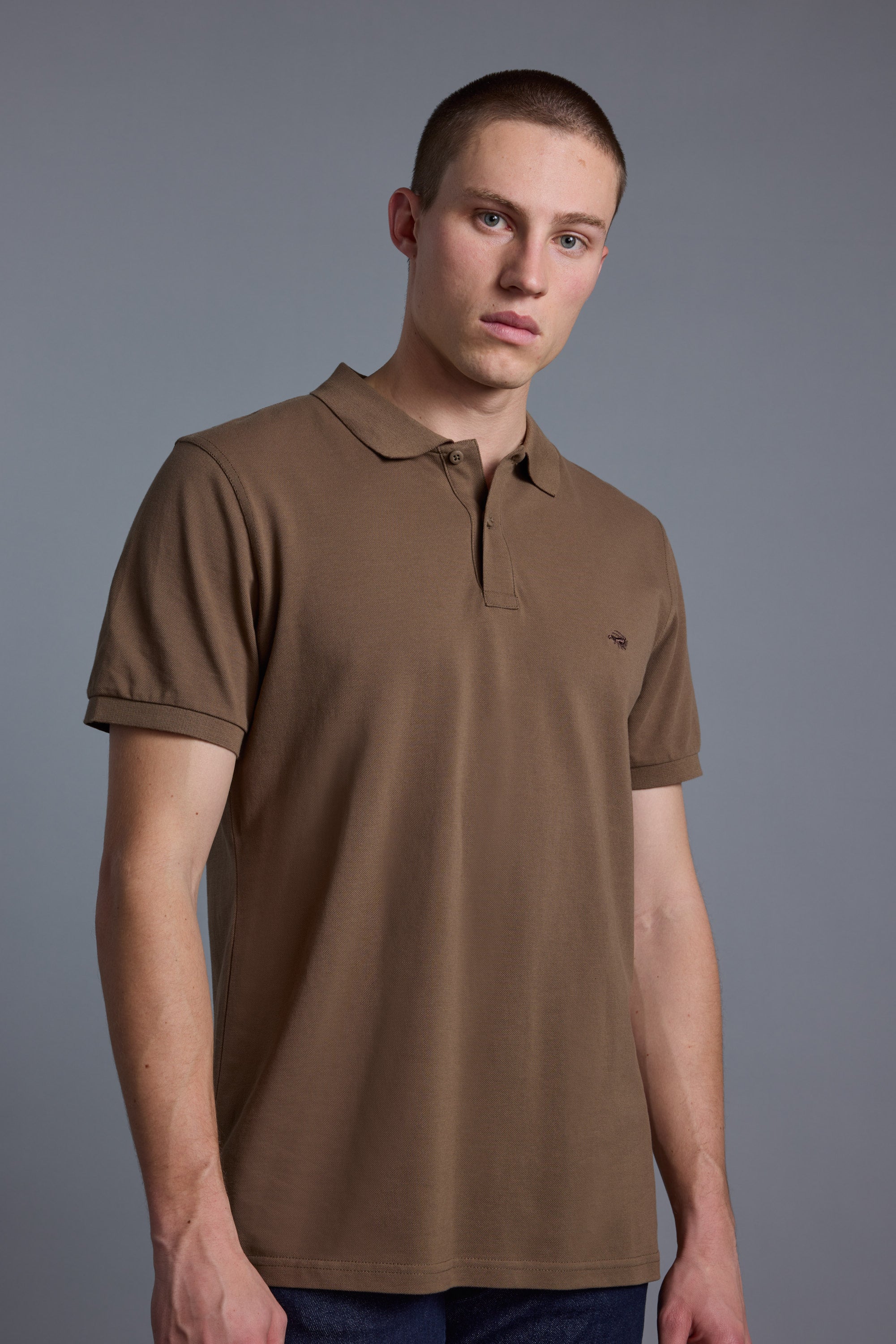 A young man with short hair, wearing a Barkers Regent Pique Polo in brown and dark jeans, stands against a plain gray background and looks calmly at the camera.