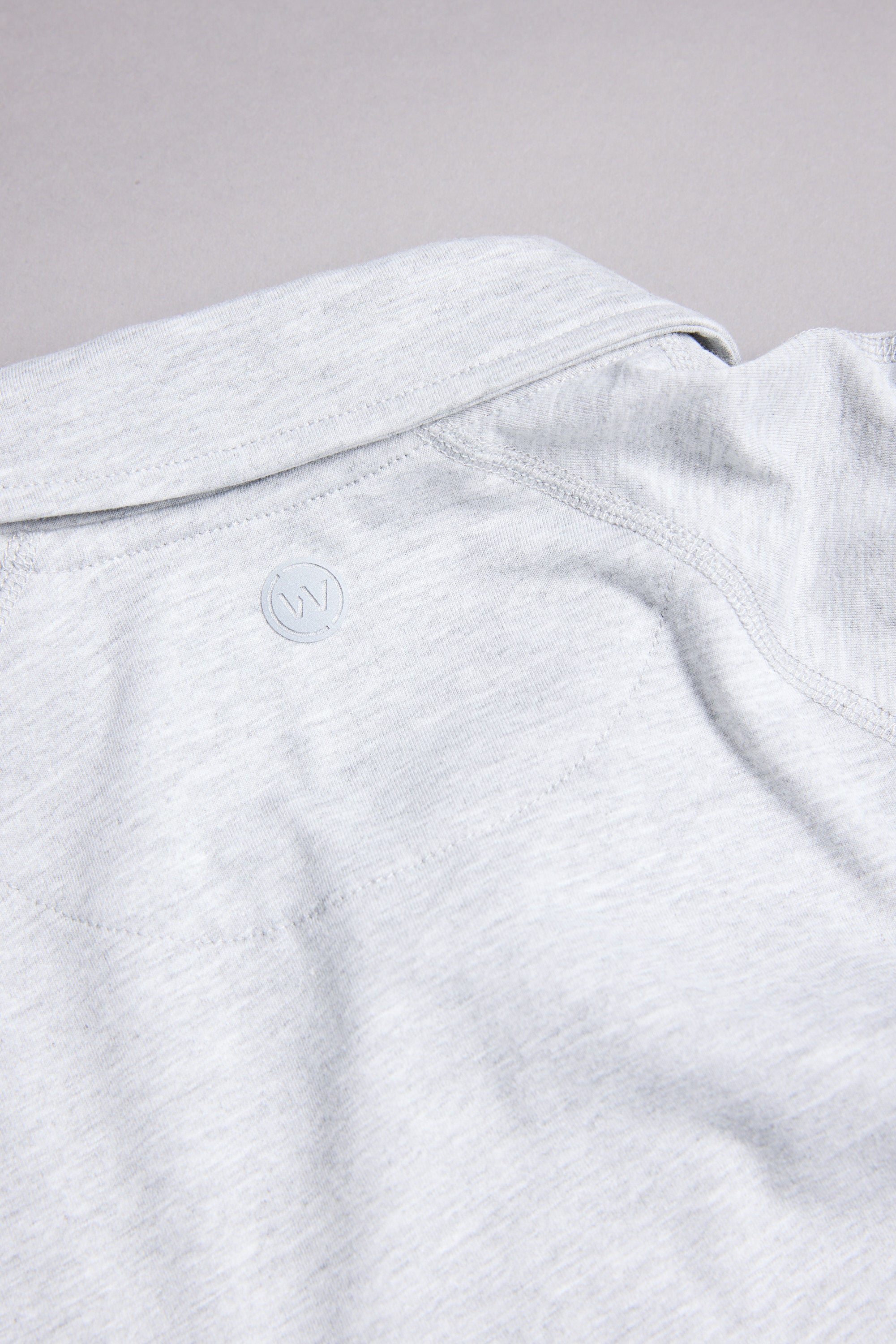 A close-up of the back collar of the WearEver Outbound Polo by WearEver in light gray, showing a subtle embossed circular W logo. The soft, slightly textured fabric is crafted from a breathable cotton-blend.