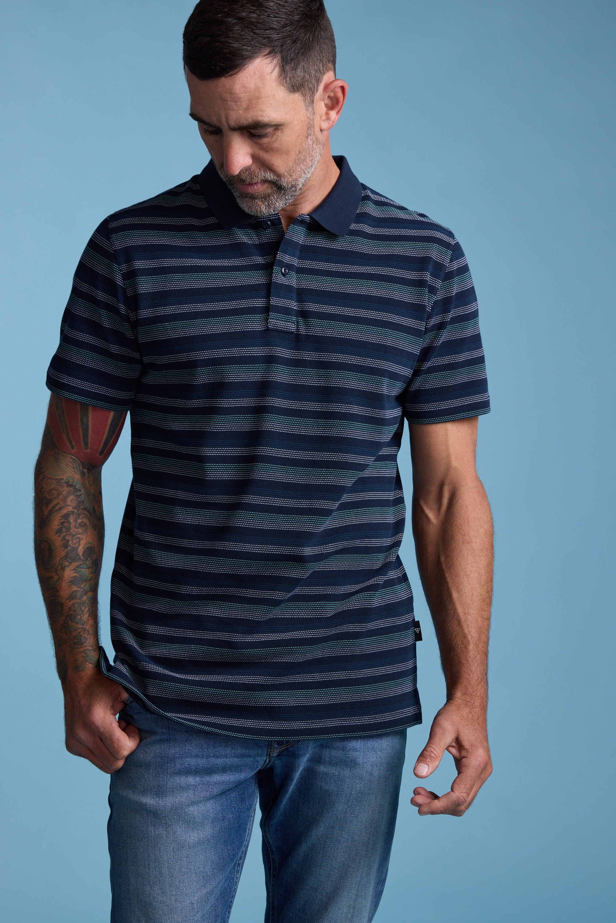 A man with short dark hair and a beard, wearing the Barkers Dalton Stripe Polo and blue jeans, stands against a light blue background, looking down with one hand on his shirt hem—a stylish summer essential.