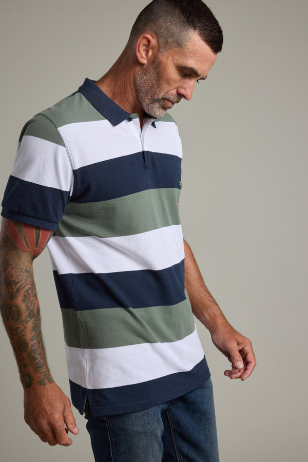 A man with short dark hair and a beard looks down, wearing the Barkers Regent Pique Stripe Polo— a limited edition design featuring navy, green, and white wide stripes. His tattooed arms rest by his sides as he pairs it with blue jeans.
