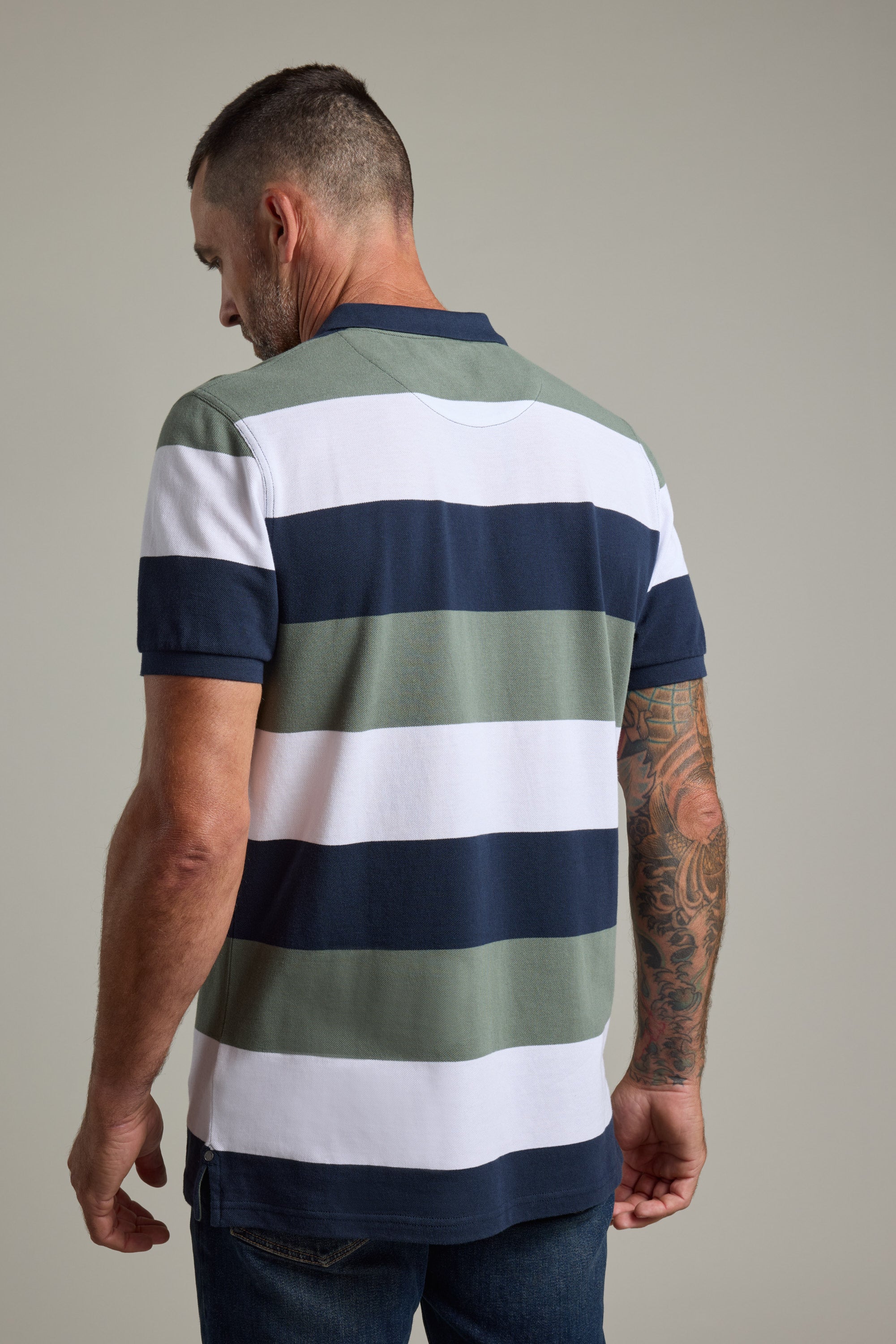A man with short hair and a tattooed right arm stands with his back to the camera, wearing the Barkers Regent Pique Stripe Polo—an organic cotton polo with wide green, navy blue, and white stripes—and dark jeans.