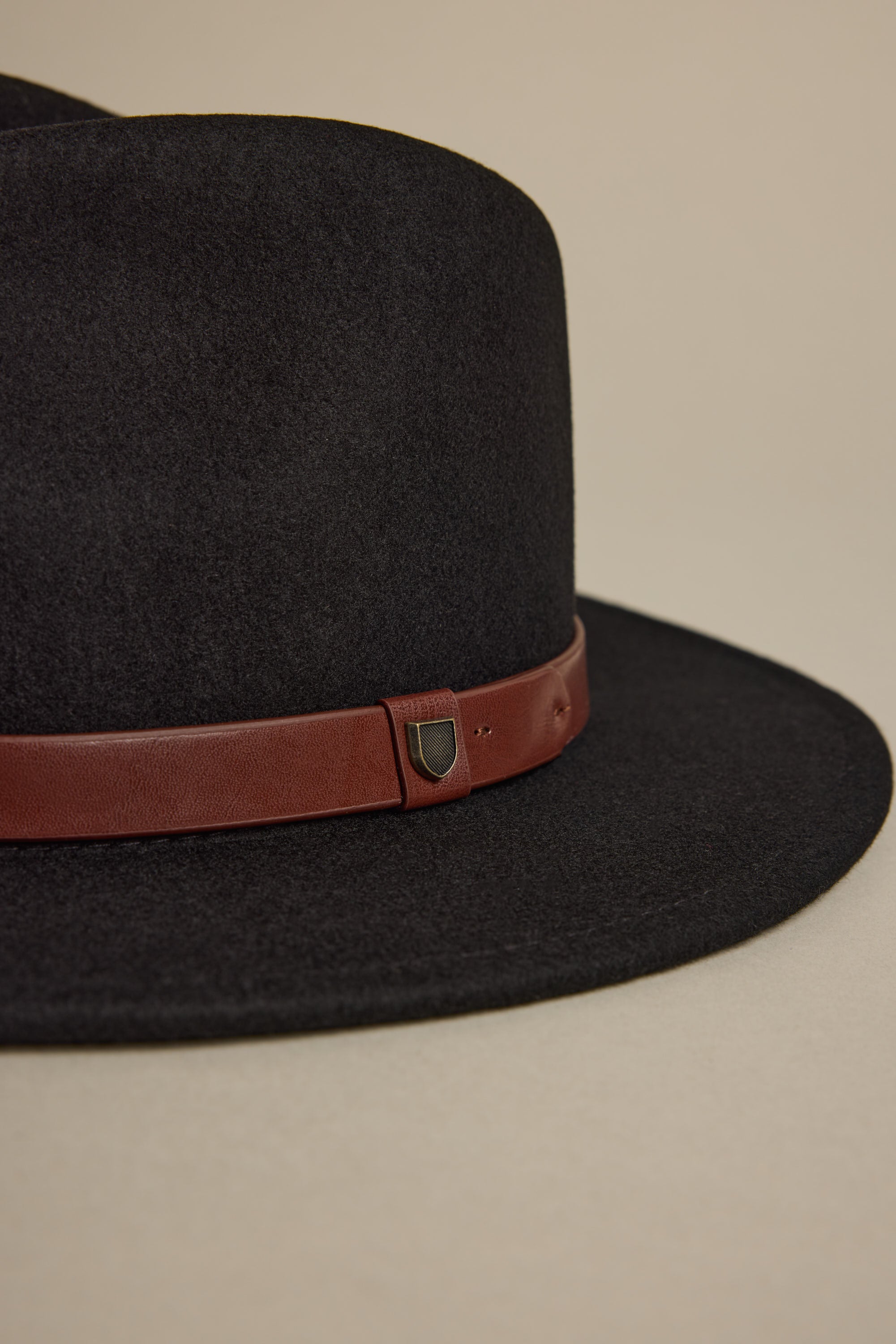A close-up of the Brixton Messer Fedora Hat—a black wool felt fedora with a wide brim and brown leather band, detailed by a small metallic shield accent. The hat is shown against a plain beige background.