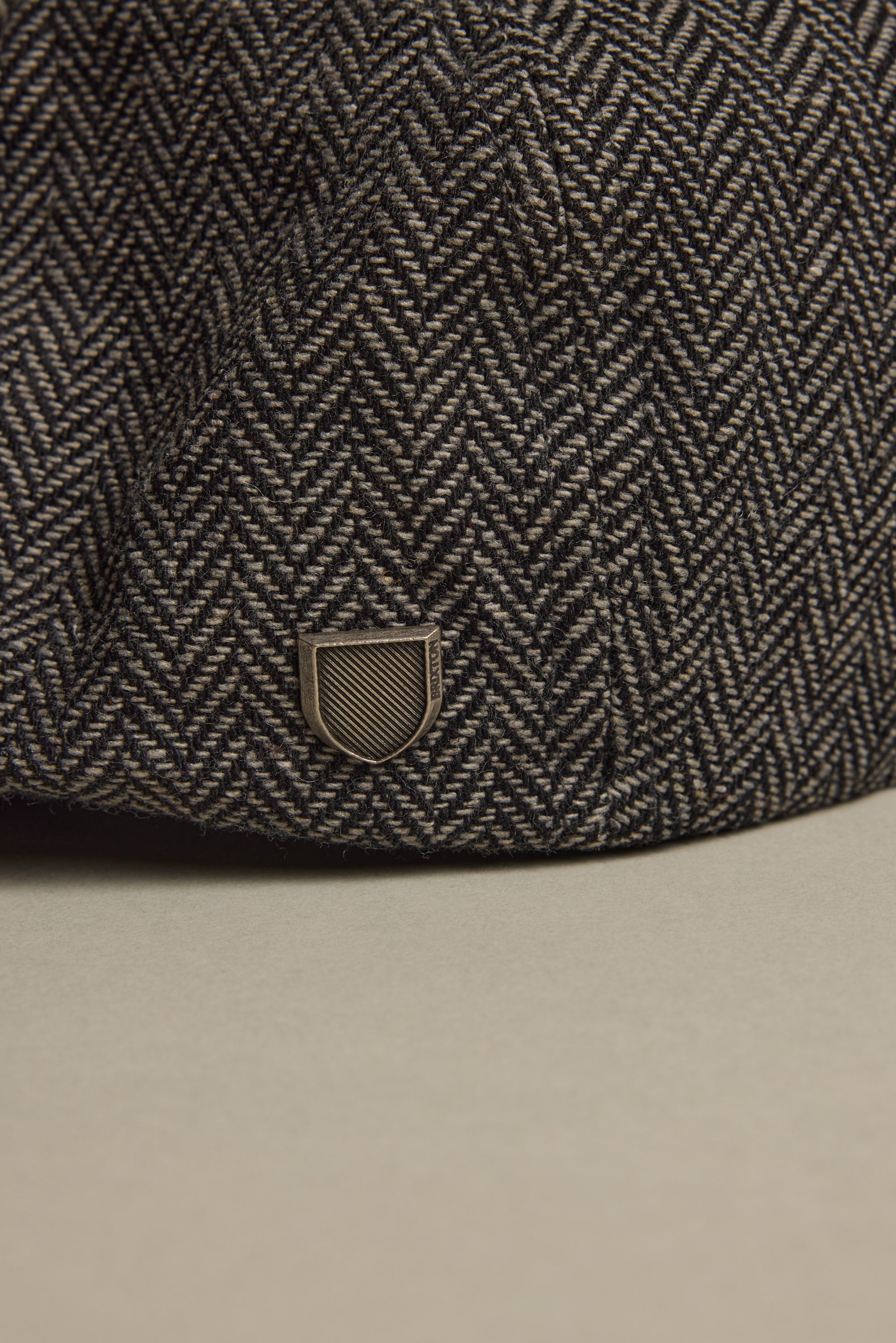 Close-up of the Brixton Brood Snap Cap by Brixton, featuring a textured herringbone wool blend and a small metallic shield emblem near the brim, set against a neutral background.