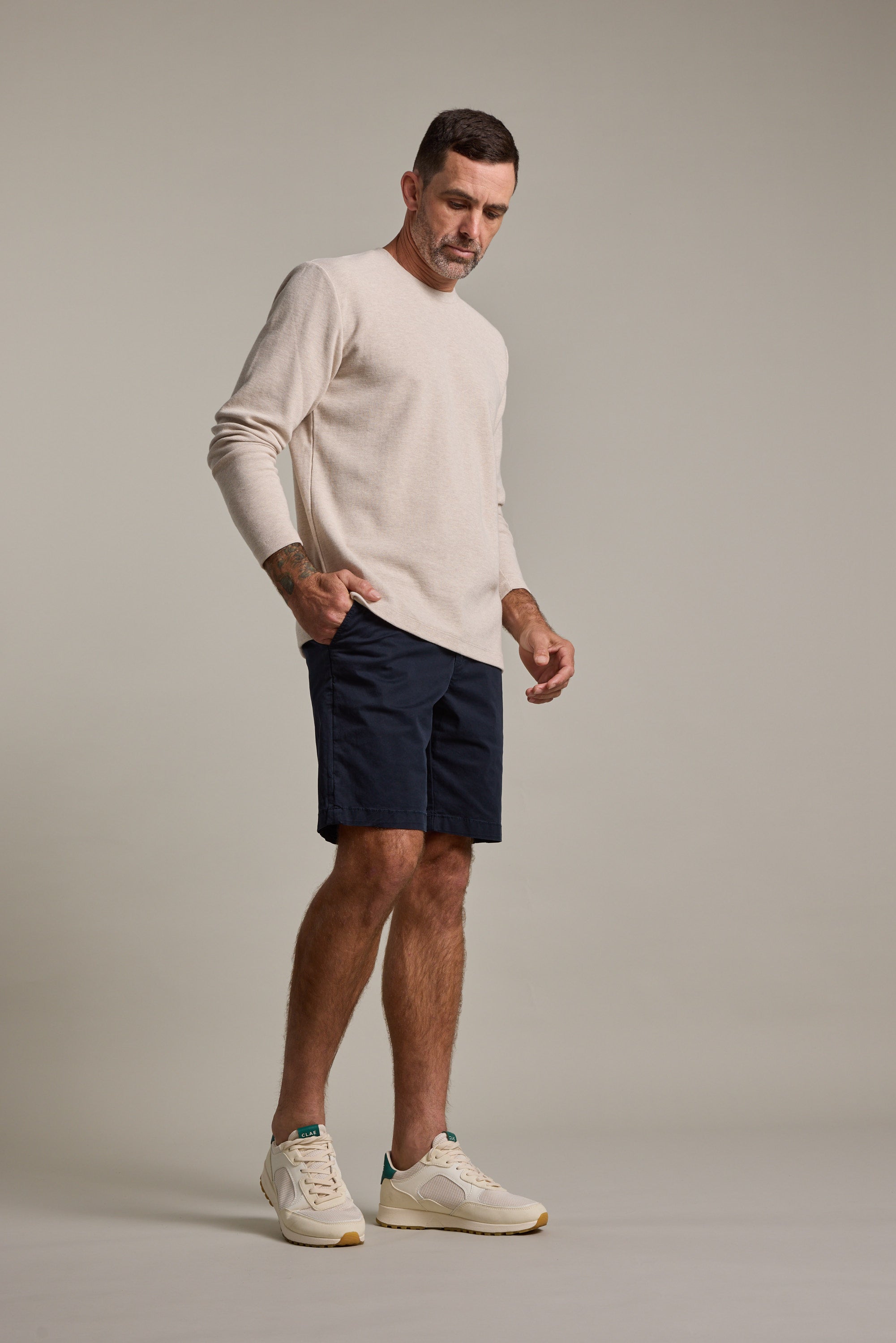 A man with short dark hair stands against a plain background wearing Barkers Kingly Chino Shorts in dark navy, a light beige long-sleeve shirt, and white sneakers, appearing to look down.
