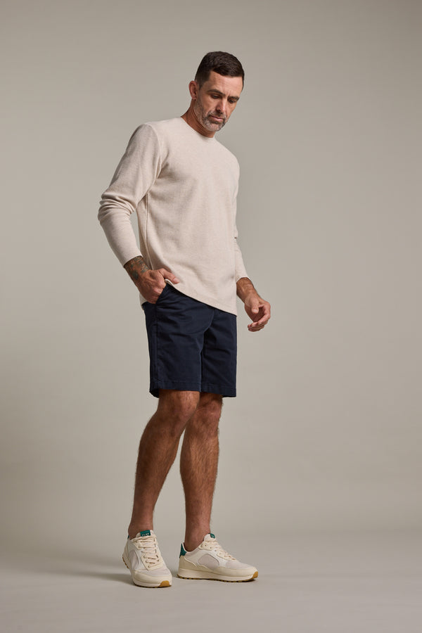 A man with short dark hair stands against a plain background wearing Barkers Kingly Chino Shorts in dark navy, a light beige long-sleeve shirt, and white sneakers, appearing to look down.