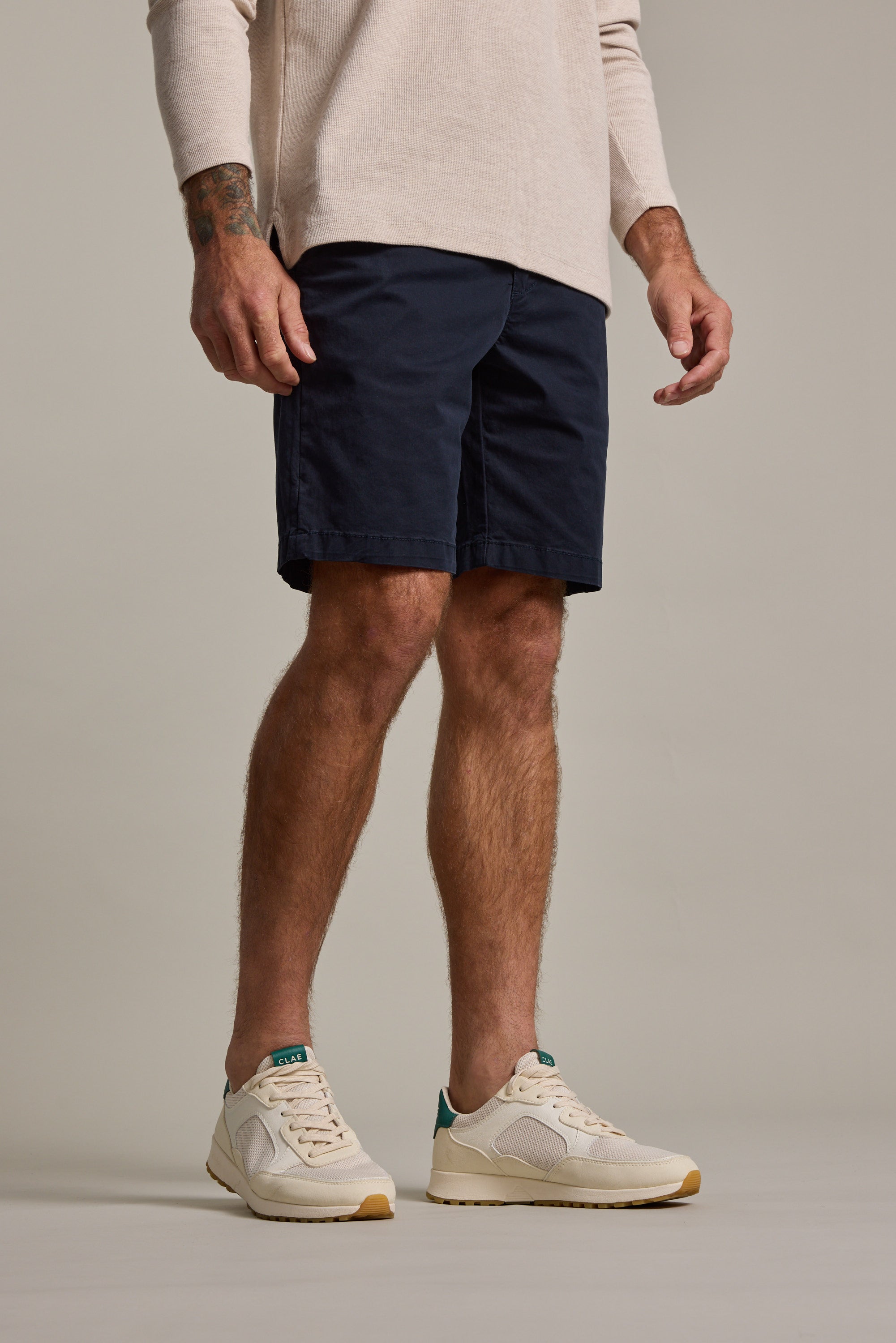 Only the lower half of a person is visible, showing them wearing Barkers Kingly Chino Short in navy, paired with a beige long-sleeve shirt and white sneakers with light brown soles against a neutral background.
