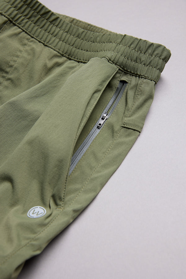 Close-up of WearEver Agile Short by WearEver: green lightweight stretch shorts with elastic waistband, zippered side pocket, and small circular logo near the hem. Shown on a light grey background. Ideal 7-inch inseam training shorts.