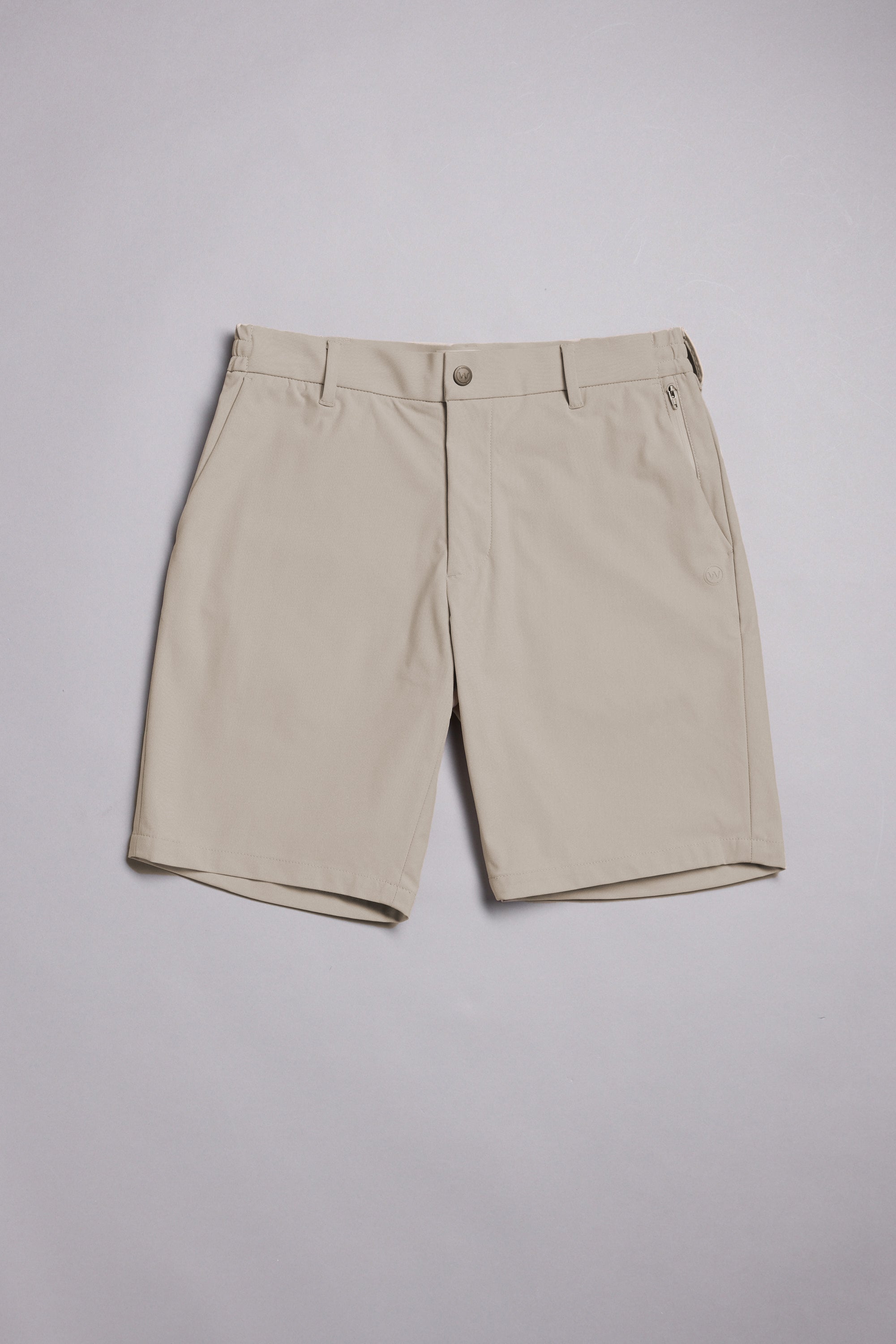 The WearEver Switch Short by WearEver is a beige, knee-length performance short with comfort stretch, belt loops, two side pockets, a zippered right pocket, and button closure, shown laid flat on a light gray background.