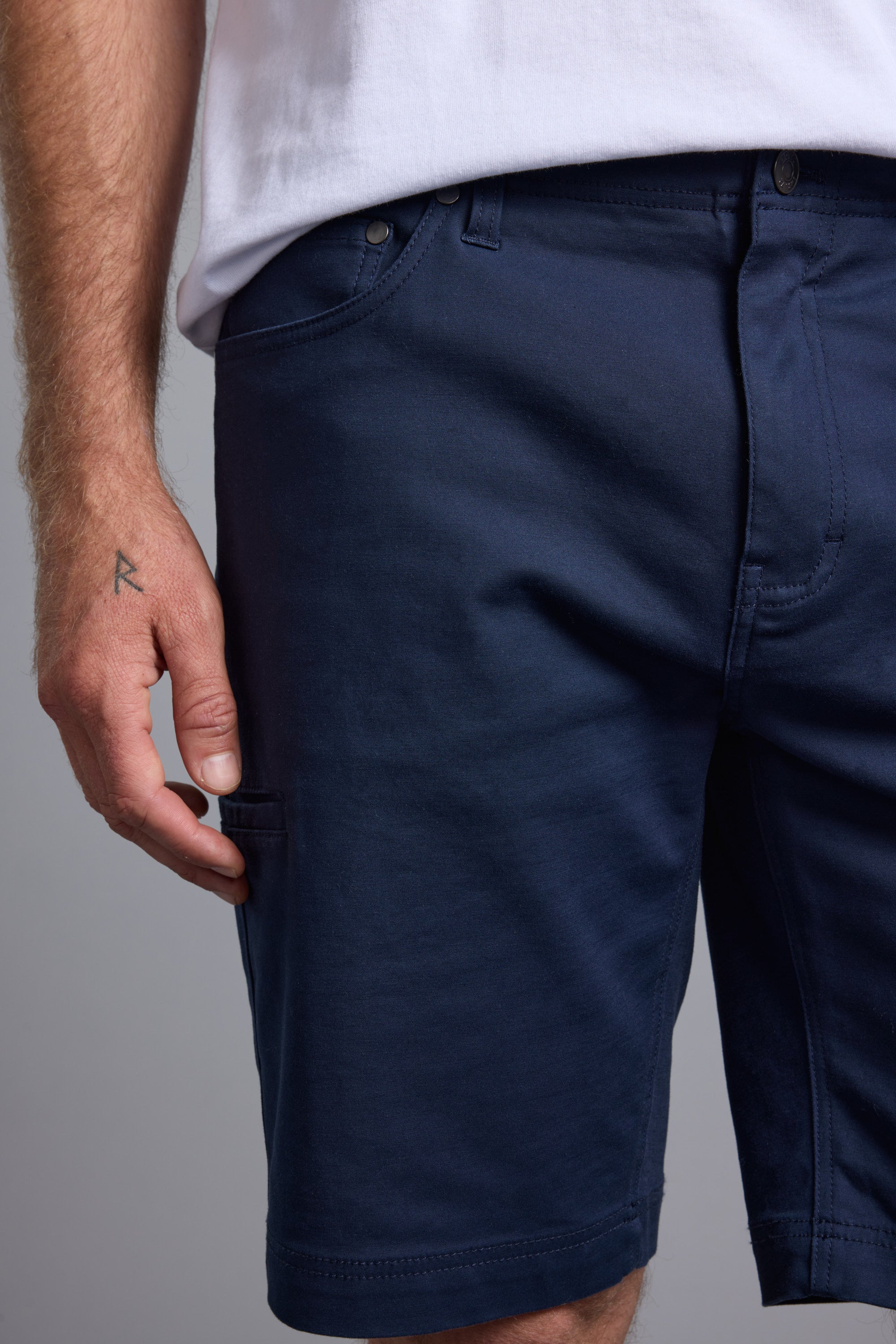 A person wearing a white T-shirt and navy Barkers Anvil 5Pkt Shorts, made from a durable cotton blend, has their left hand (with an R tattoo) resting by their side.
