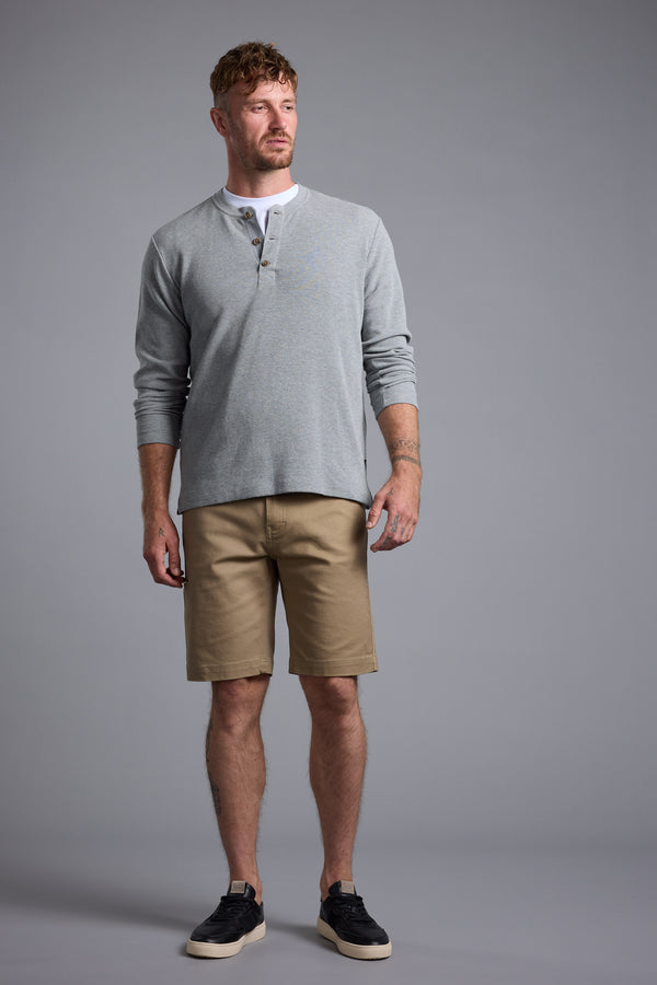 A man with short hair and a beard stands against a gray background, wearing a light gray long-sleeve henley shirt, Barkers Anvil 5Pkt Short in tan with an elasticated waistband, and black sneakers with white soles. He looks slightly left, expression neutral.