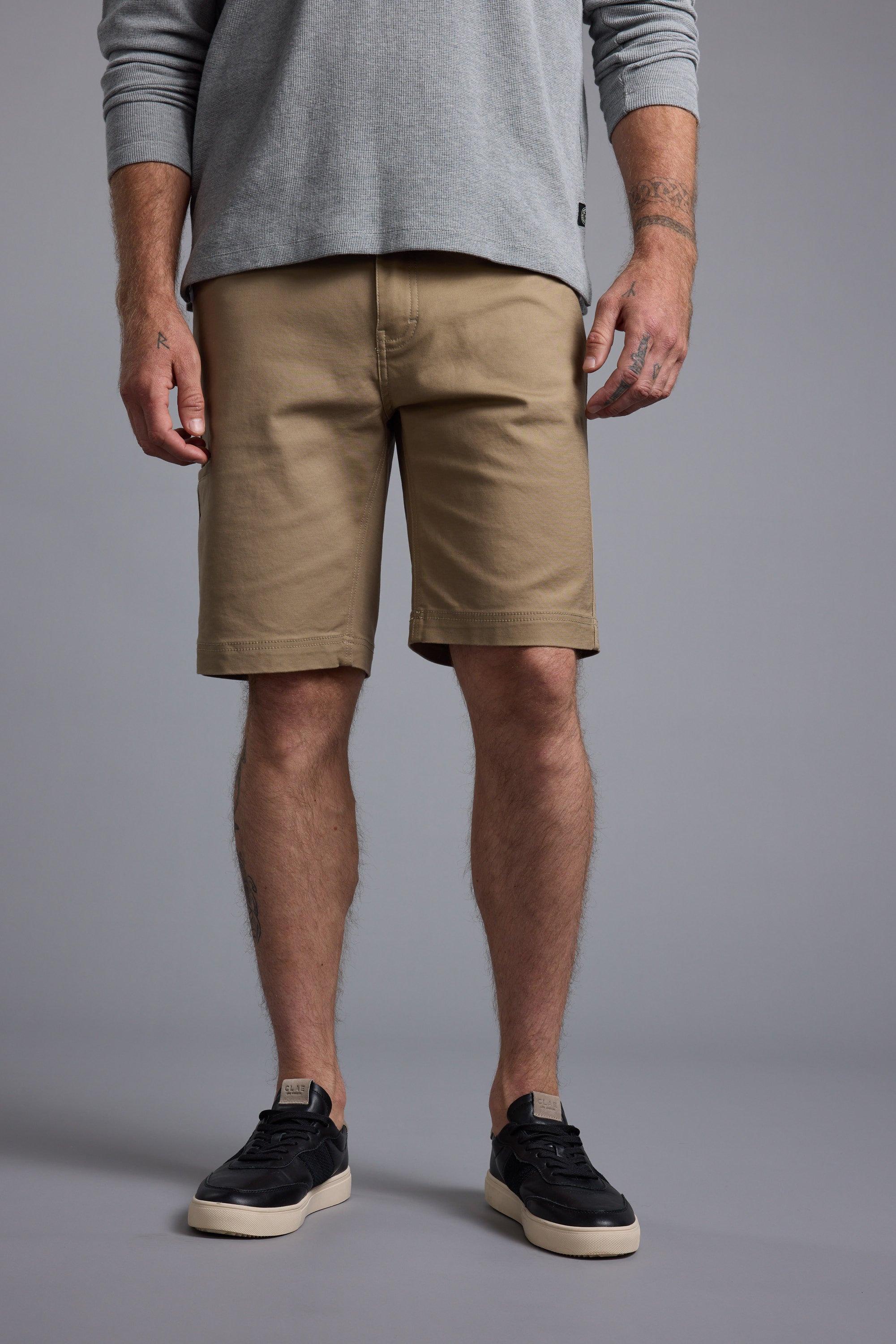 A person, visible from chest to feet, wears a light gray long-sleeve shirt, Barkers Anvil 5Pkt Short with an elasticated waistband, and black sneakers with white soles against a plain gray background.