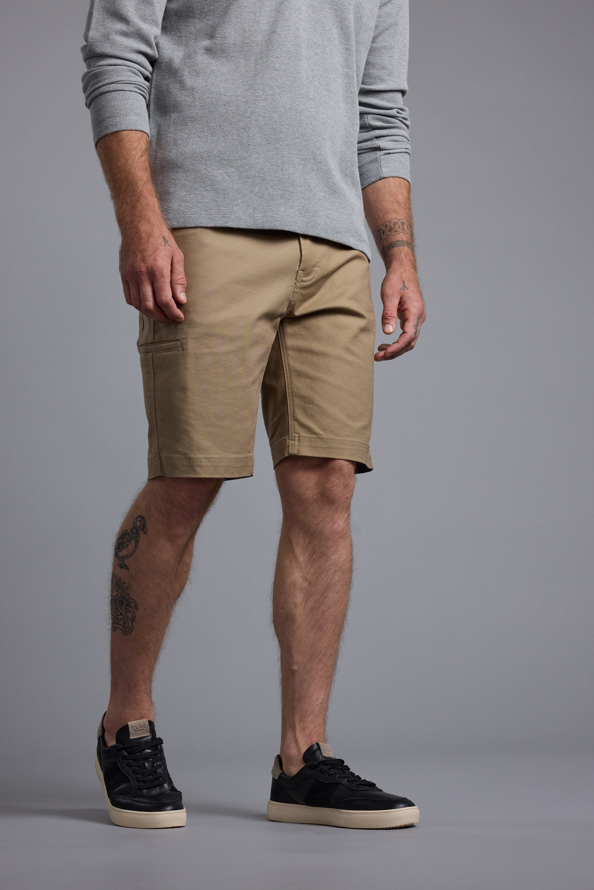 A man wearing a light gray long-sleeve shirt, Barkers Anvil 5Pkt Shorts in tan with an elastic waistband, and black sneakers with white soles stands against a plain gray background. Tattoos are visible on his right forearm and left calf; face not shown.
