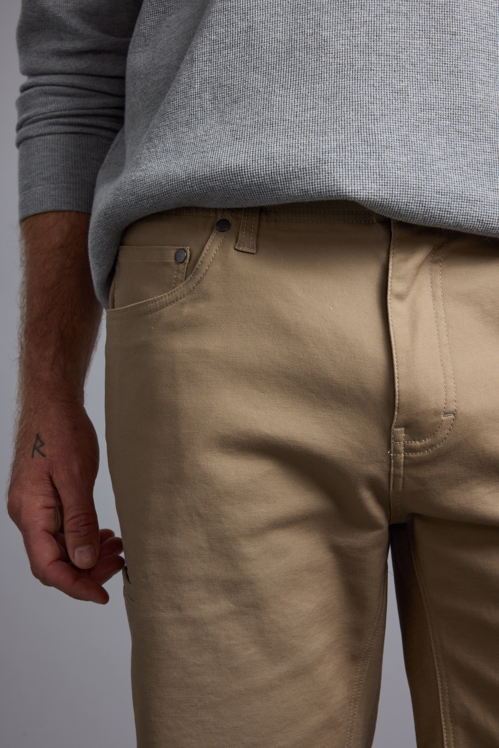 A person wears a light gray sweater tucked into beige Barkers Anvil 5Pkt Short with an elasticated waistband. Their left hand, featuring a small "R" tattoo, is visible. The photo is cropped at the chest and mid-thigh.
