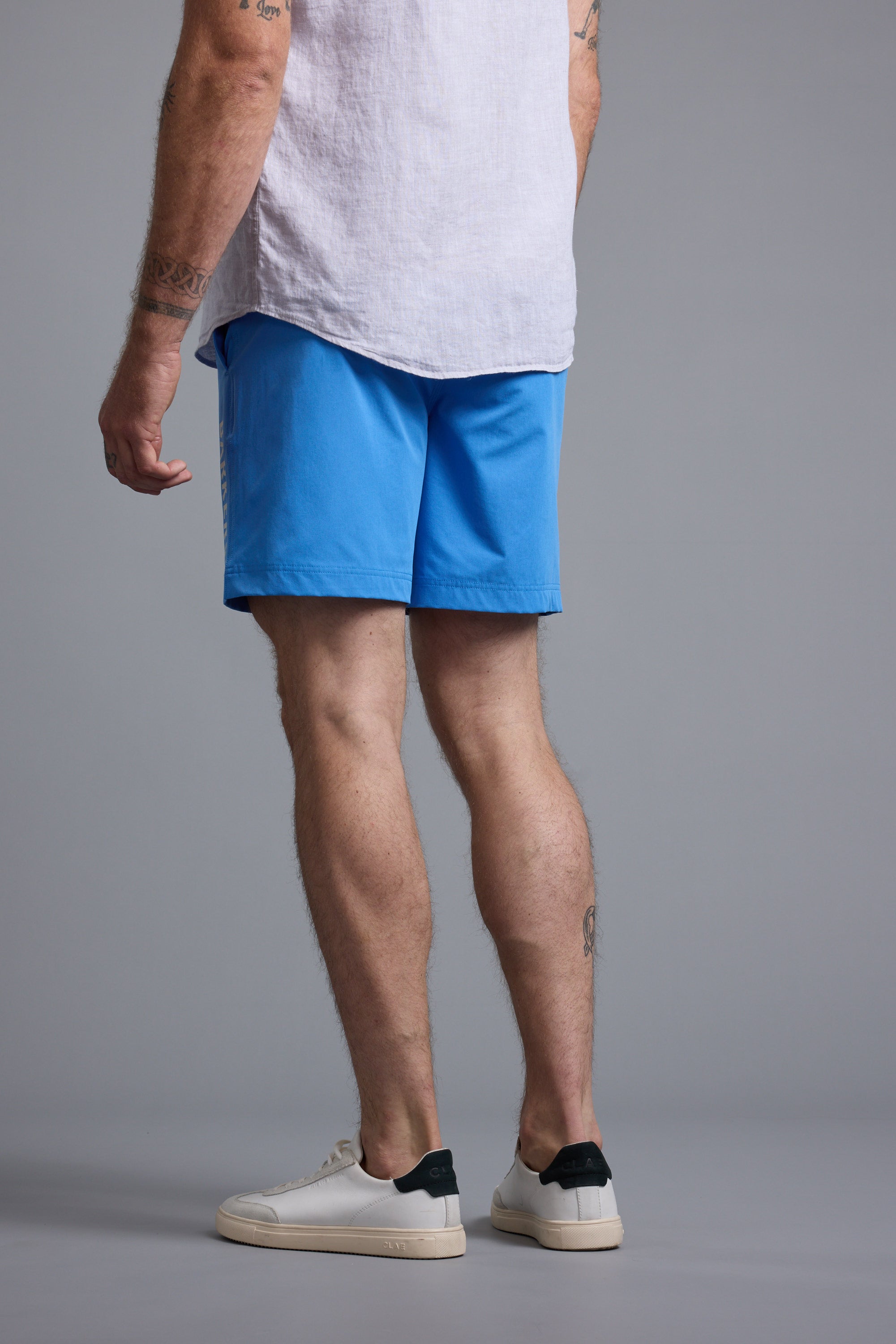 A man stands with his back to the camera, wearing a light shirt and bright blue Barkers Weekend Shorts by Barkers, made from recycled polyester, paired with white sneakers featuring dark heel accents against a plain gray background.