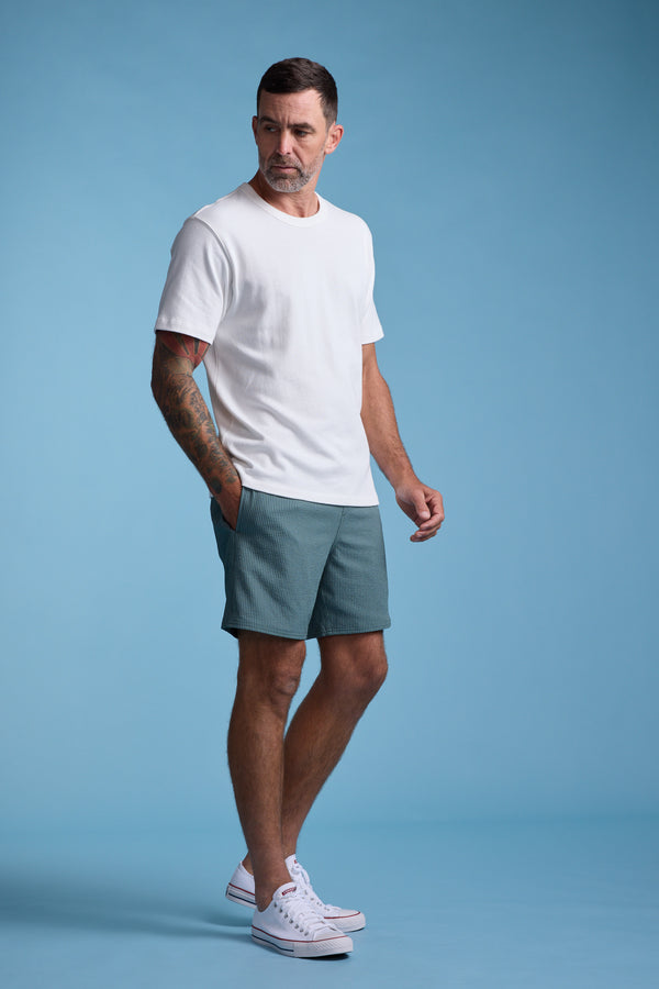 A man with short hair and arm tattoos wears a white tee, Barkers Dehaven Stripe Volley quick-dry swim trunks in green, and white sneakers, standing against a plain light blue background.
