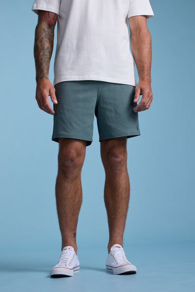 A man stands against a blue background wearing a white t-shirt, Barkers Dehaven Stripe Volley shorts, and white sneakers. His arms are relaxed at his sides, showing tattoos on his left arm. His head is not visible in the image.