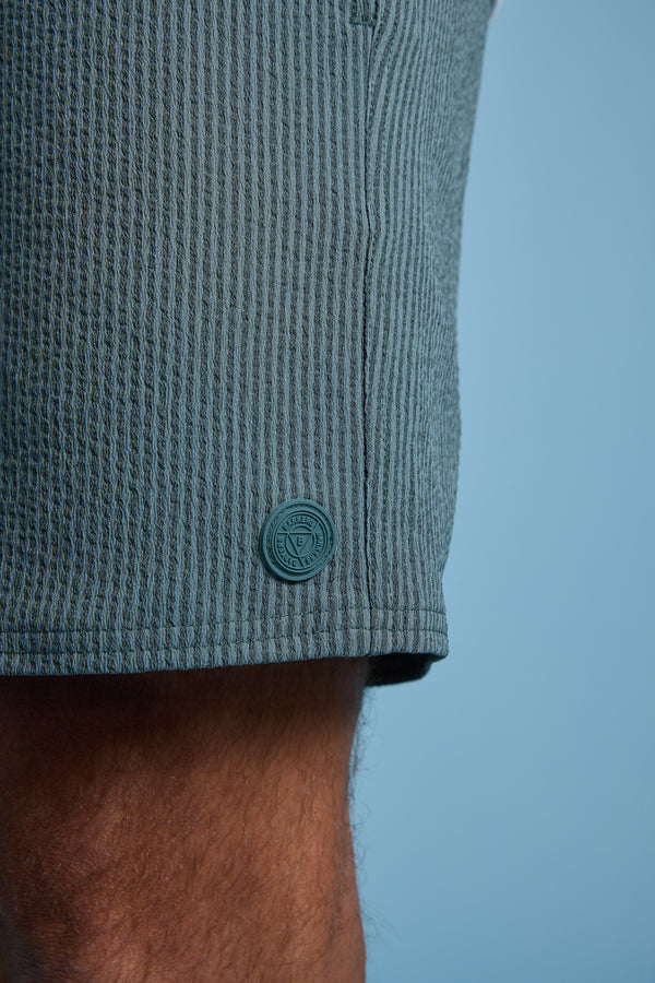 Close-up of a person's upper leg wearing Barkers Dehaven Stripe Volley shorts in textured, light greenish-blue seersucker with a matching round button and small emblem by the hem, set against a plain light blue background.