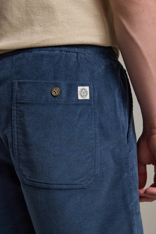 A close-up of someone wearing blue Barkers Montauk Cord Weekend Shorts with an elastic waistband, buttoned back pocket, and a small white label by the pocket. The person’s arm and a beige shirt are partially shown.