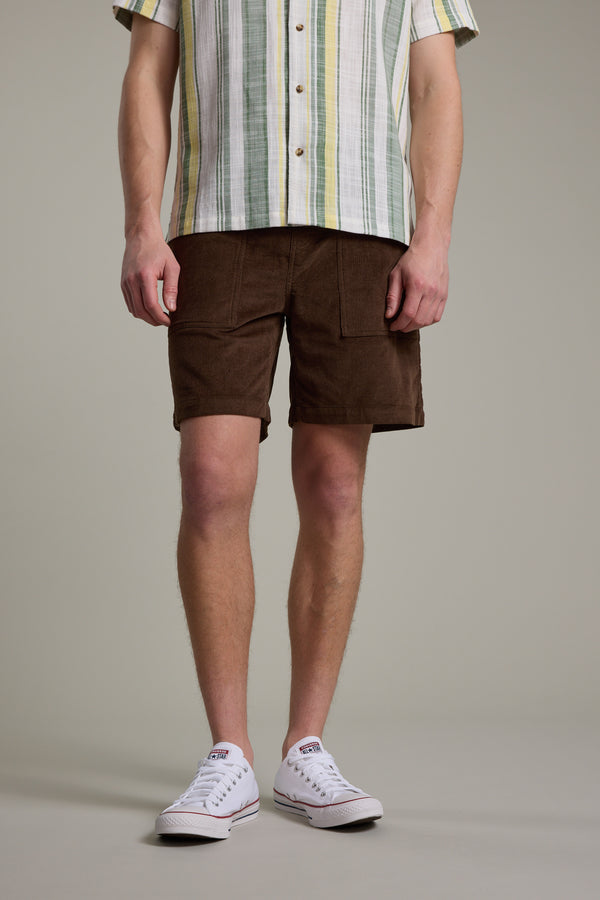 A person wearing a short-sleeved, striped button-up shirt, Barkers Montauk Cord Weekend Short with an elastic waistband, and white low-top Converse sneakers stands against a plain background. Their face is not visible.