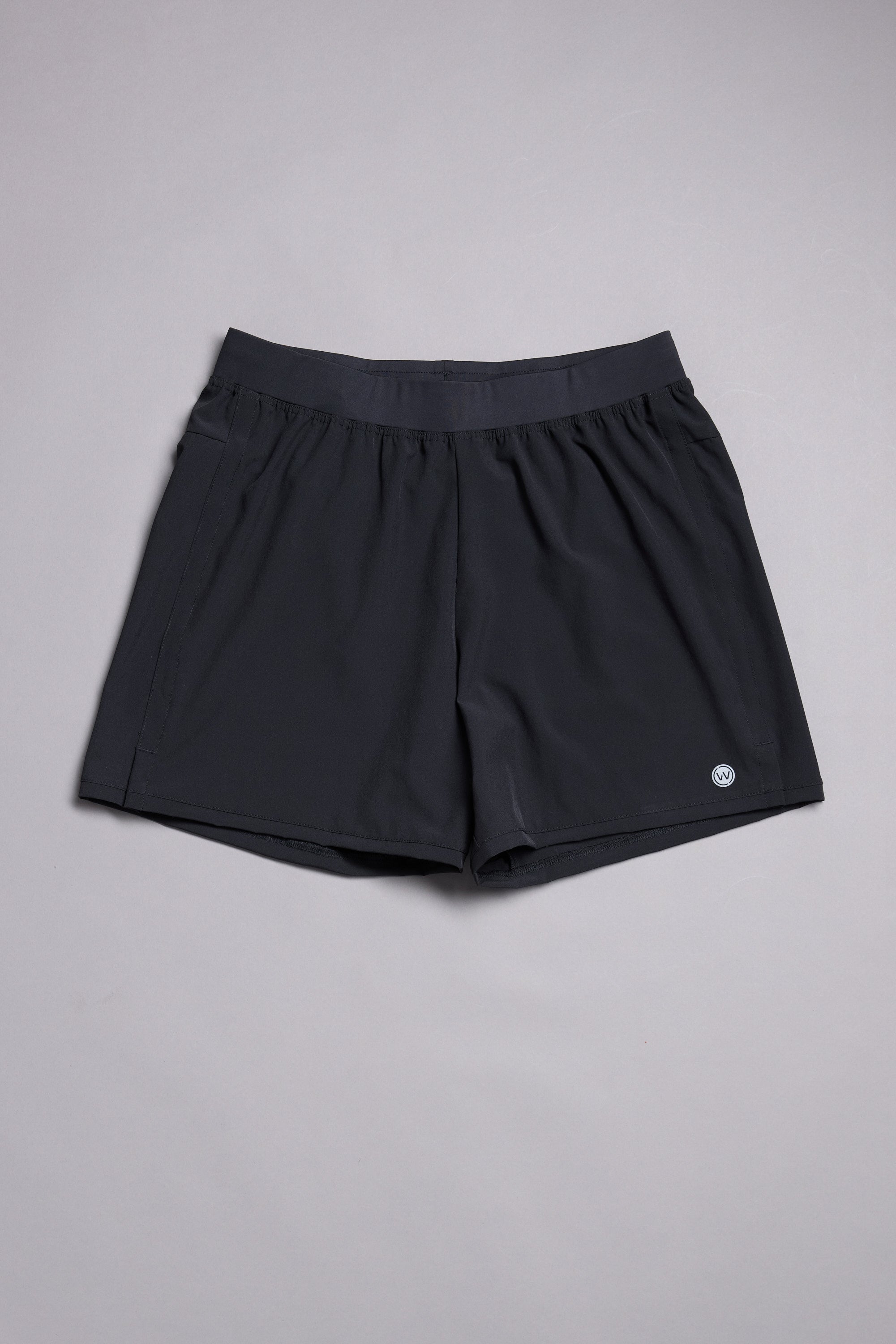 The WearEver Cadence Short, a pair of lightweight black workout shorts with a 5-inch inseam and elastic waistband, is laid flat on a light gray surface. A small round WearEver logo appears near the bottom hem on one leg.