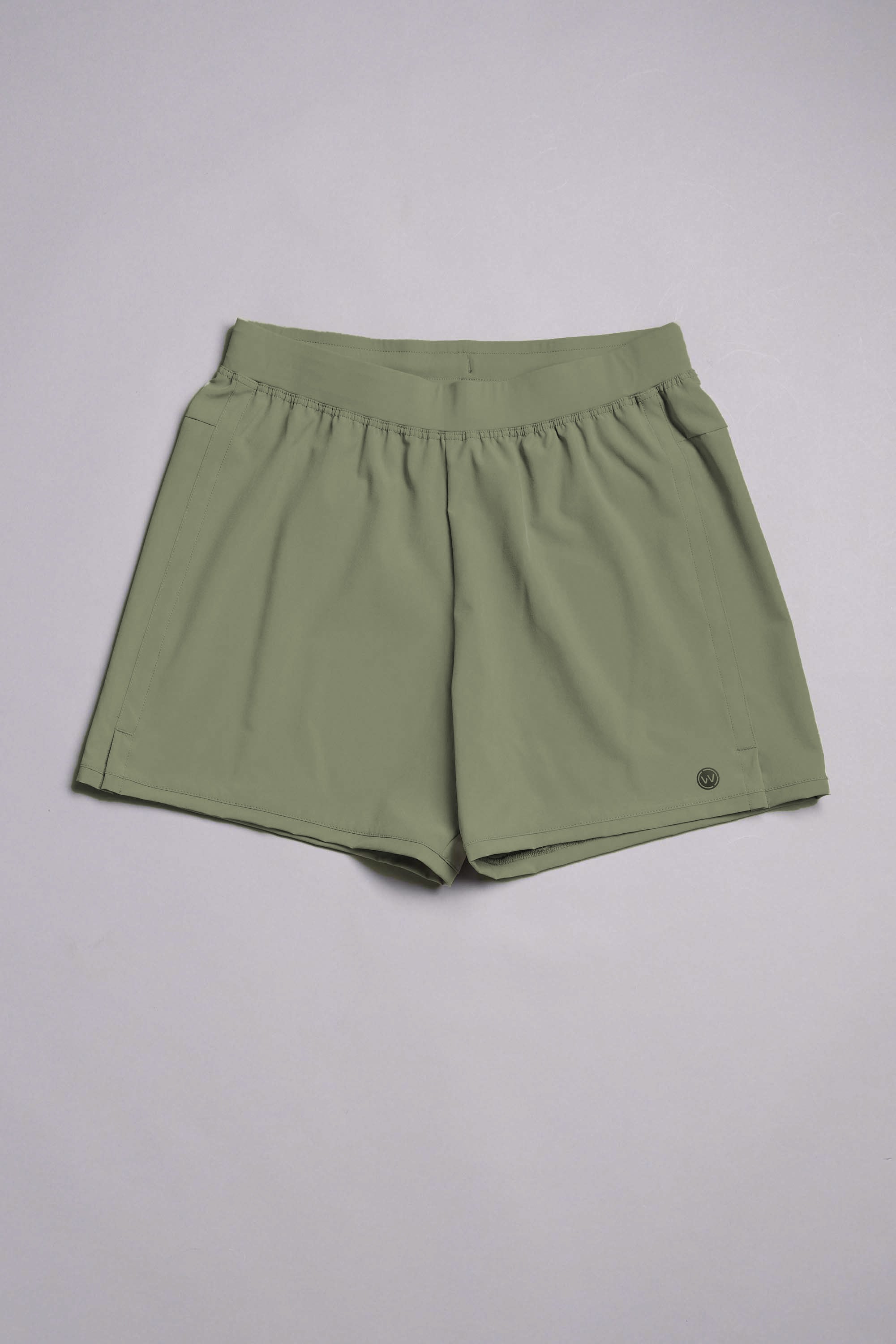 The WearEver Cadence Short in light olive green features a 5-inch inseam, elastic waistband, side pockets, and a small circular logo by the left hem. Made from lightweight, breathable fabric and shown flat on a light gray background.