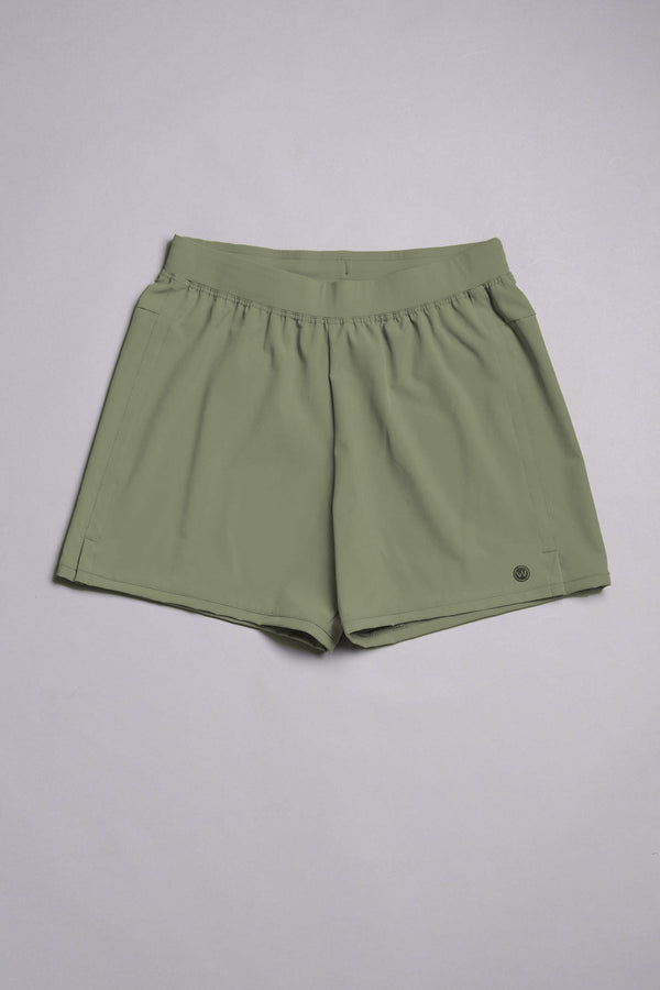 The WearEver Cadence Short in light olive green features a 5-inch inseam, elastic waistband, side pockets, and a small circular logo by the left hem. Made from lightweight, breathable fabric and shown flat on a light gray background.