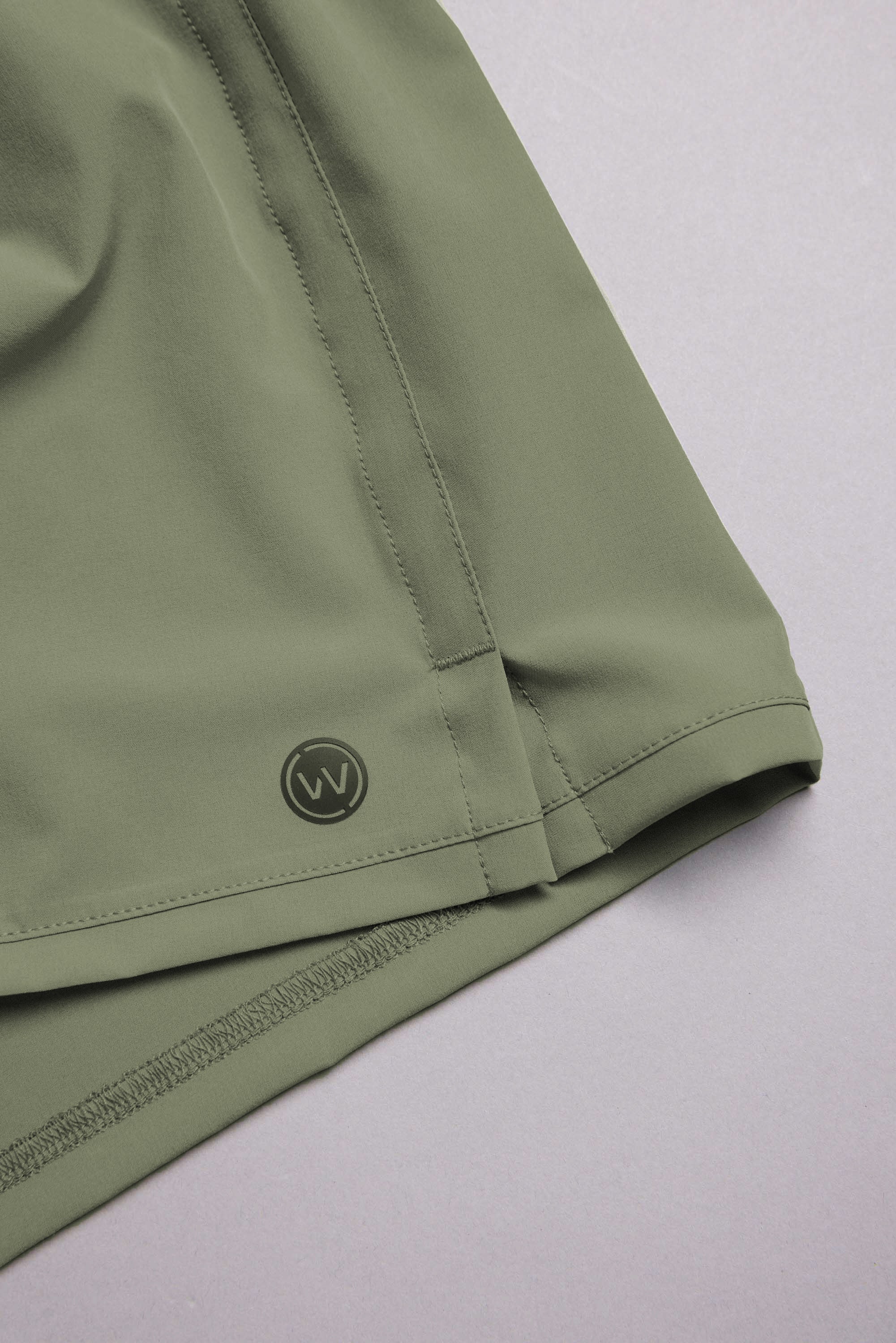 Close-up of the hem and side seam of an olive green WearEver Cadence Short with a small circular logo and 5-inch inseam, shown on a light grey background.