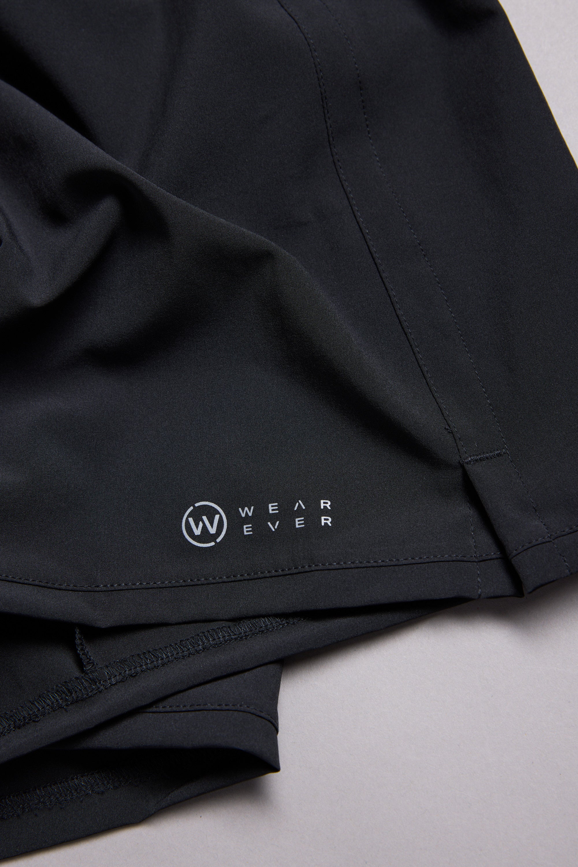 Close-up of black WearEver Cadence Shorts, featuring a 5-inch inseam and the white WearEver logo near the hem, laid flat on a light gray surface.