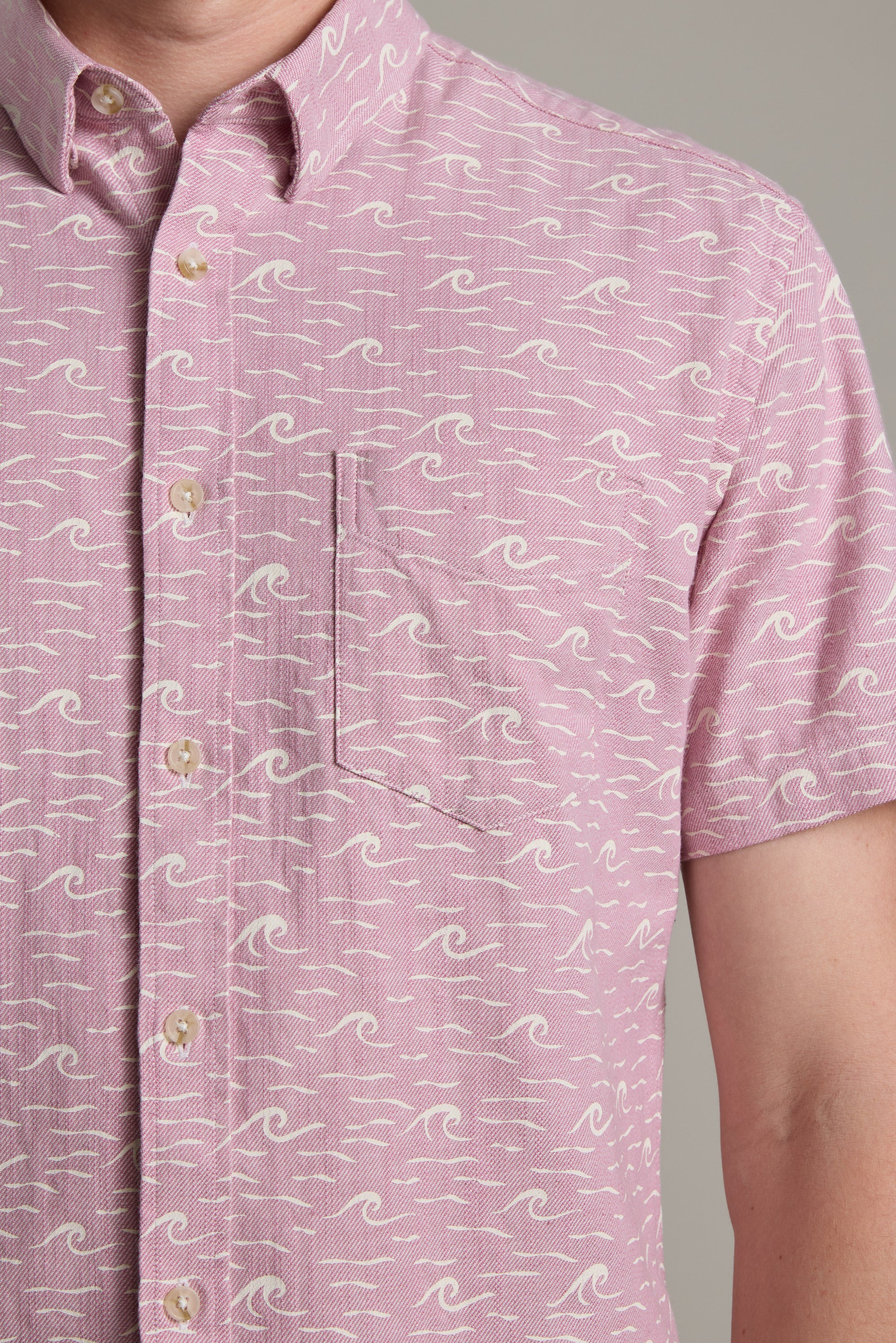 A person wearing the Barkers Shelby Print Shirt—a light pink, short-sleeve summer shirt with a white wave pattern, beige buttons, and a left chest pocket. Made from responsibly sourced cotton. Only the upper torso is visible.