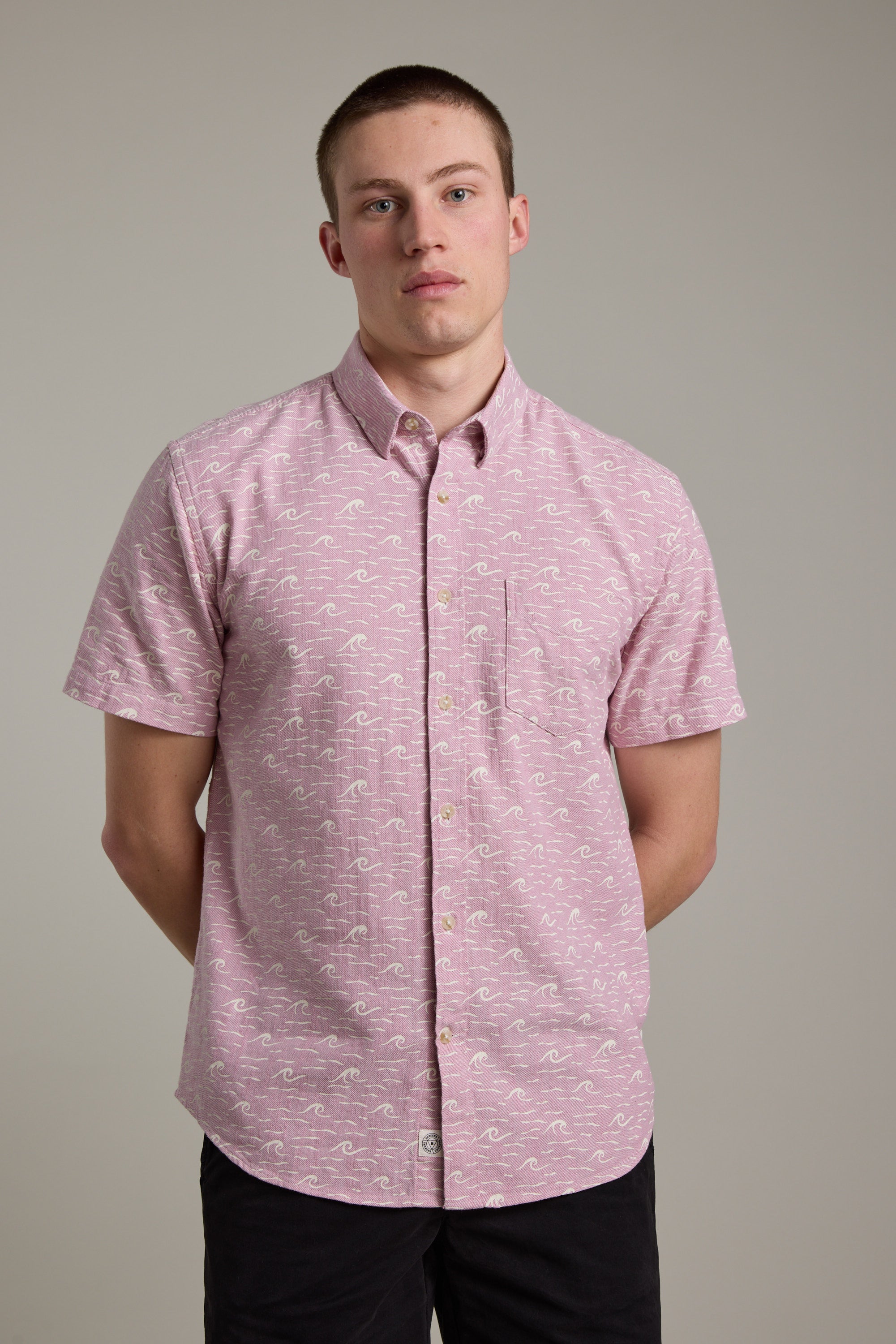A young man with short hair wears the Barkers Shelby Print Shirt, a short sleeve summer shirt made from responsibly sourced cotton. He stands against a plain background with his hands behind his back, looking neutrally at the camera.