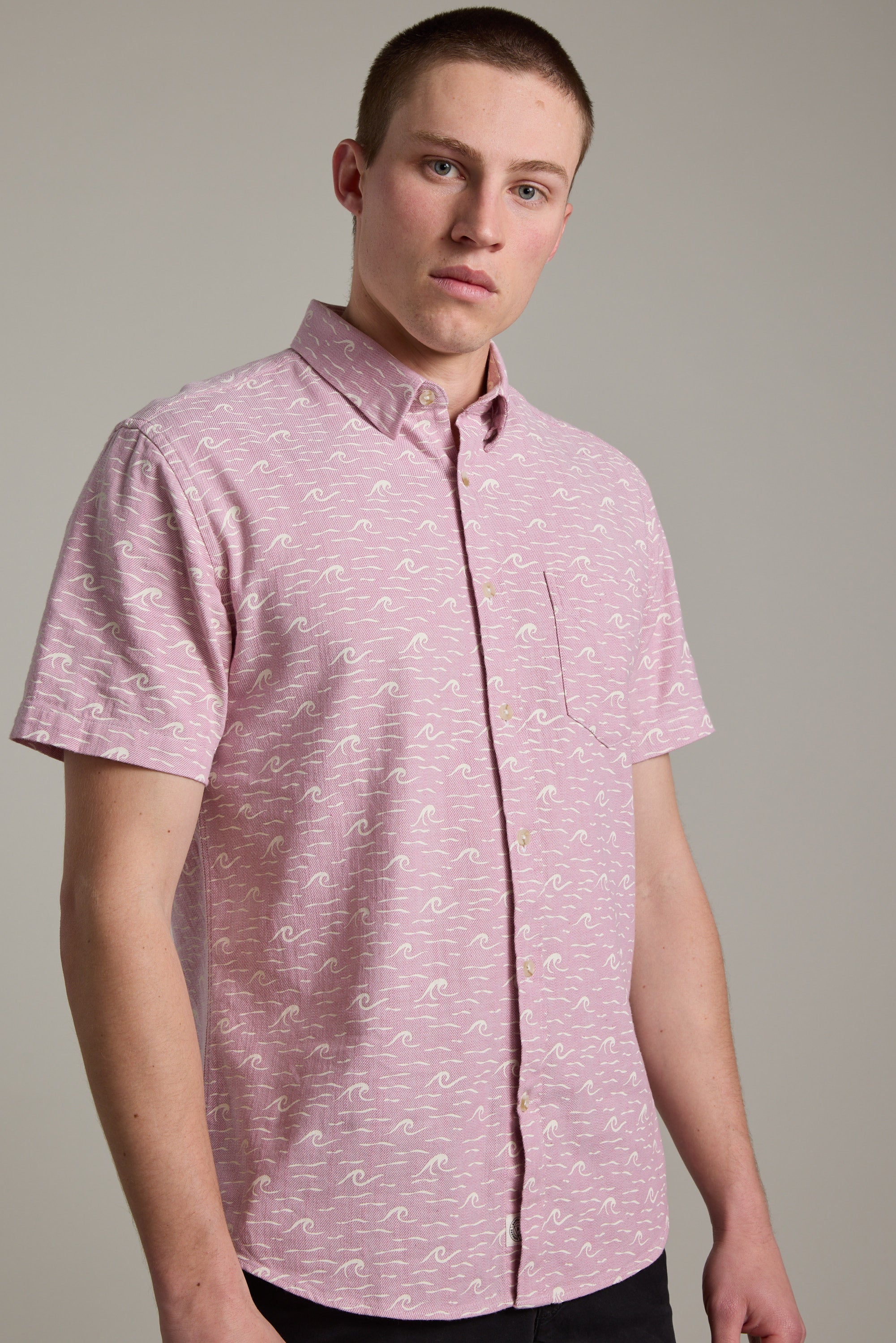 A young man wearing the Barkers Shelby Print Shirt—a short-sleeve summer shirt in responsibly sourced cotton with a subtle white wave design—stands before a plain background, gazing at the camera with a neutral expression.