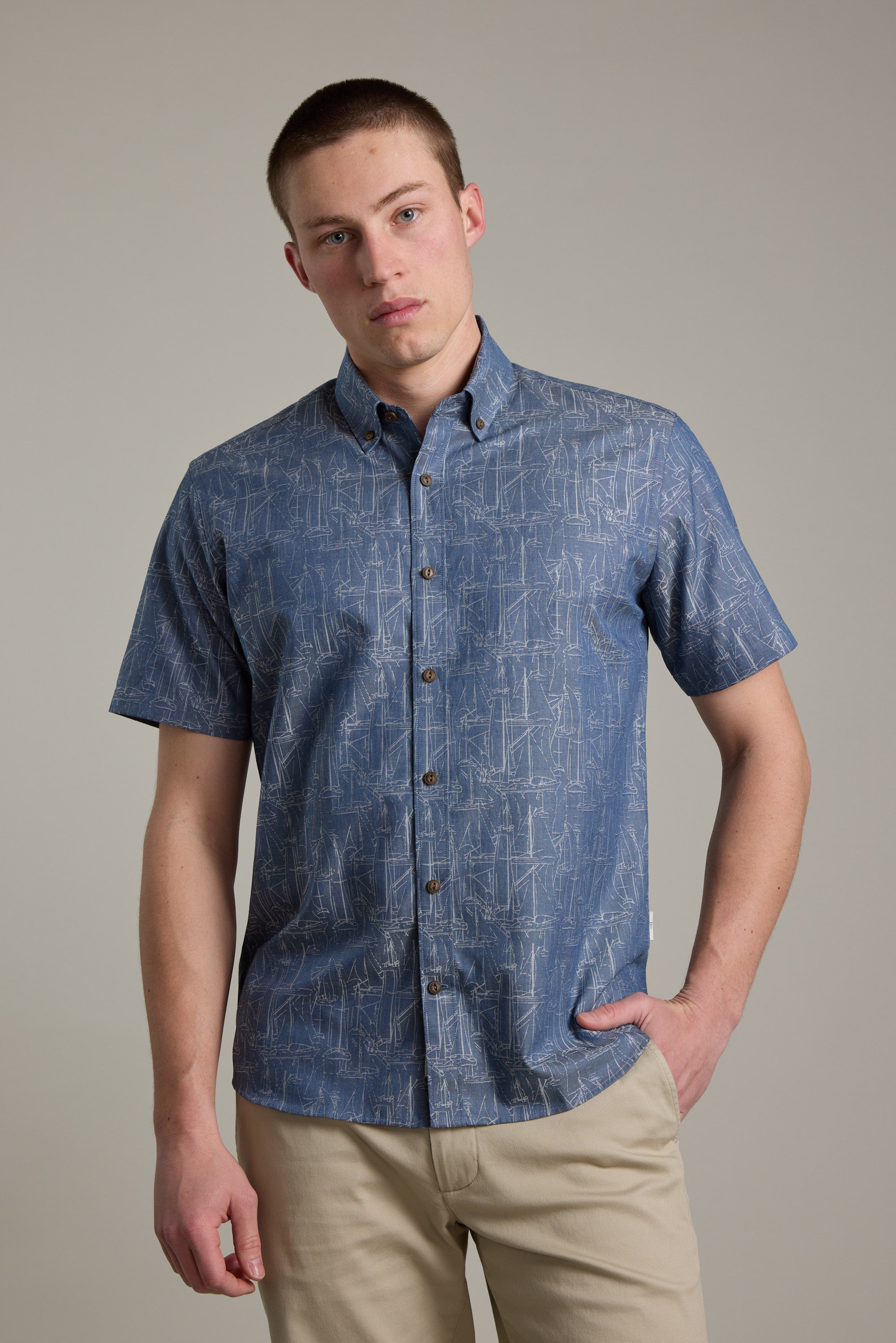 A young man models the Barkers Yacht Jacquard Shirt, a short-sleeved blue summer top, paired with beige pants against a light gray background, one hand in his pocket and wearing a neutral expression.