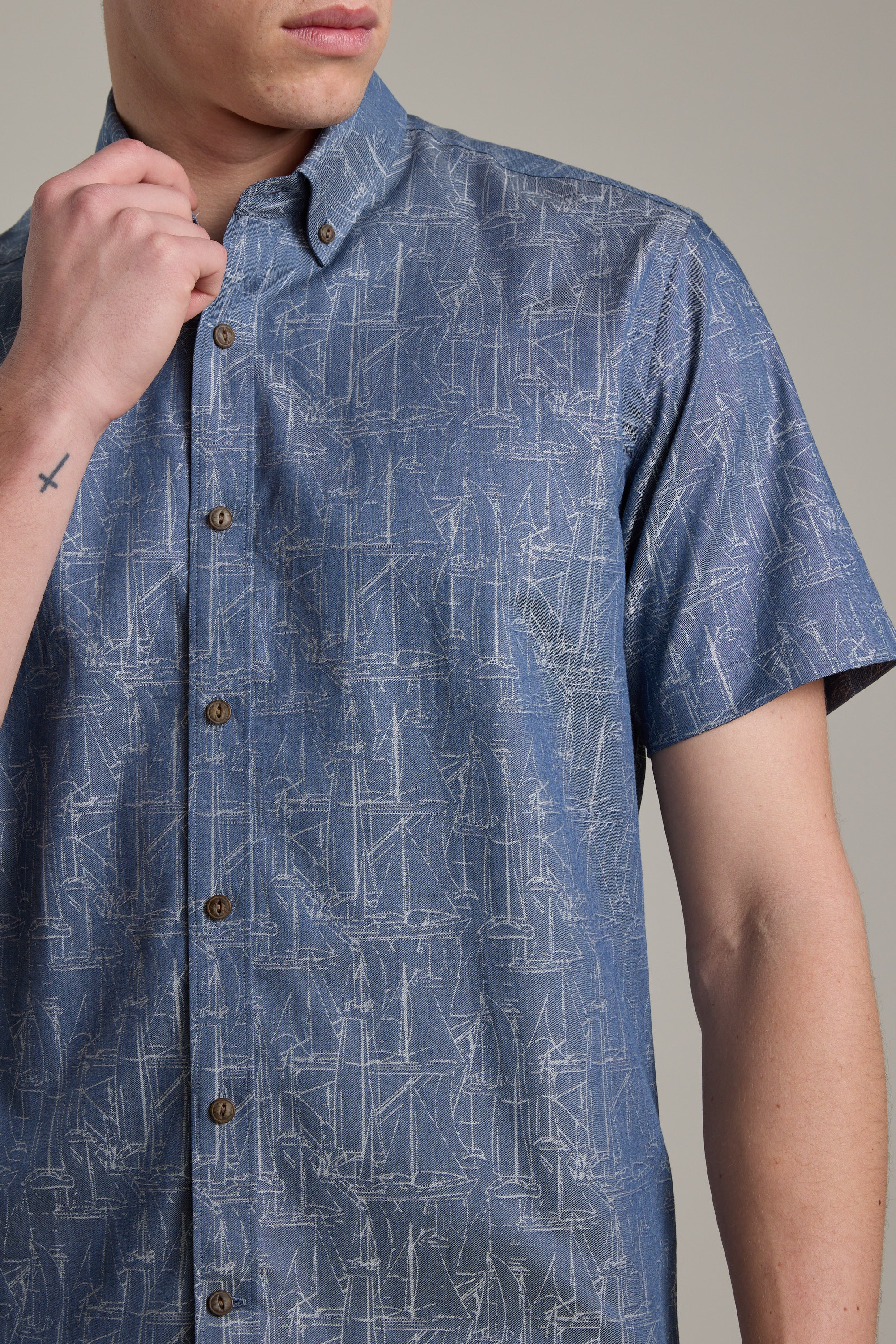 A man models the Barkers Yacht Jacquard Shirt, a blue short sleeve cotton shirt with a subtle yacht print. His left hand, showing a small cross tattoo on his wrist, is near his chin. The background is plain and neutral.