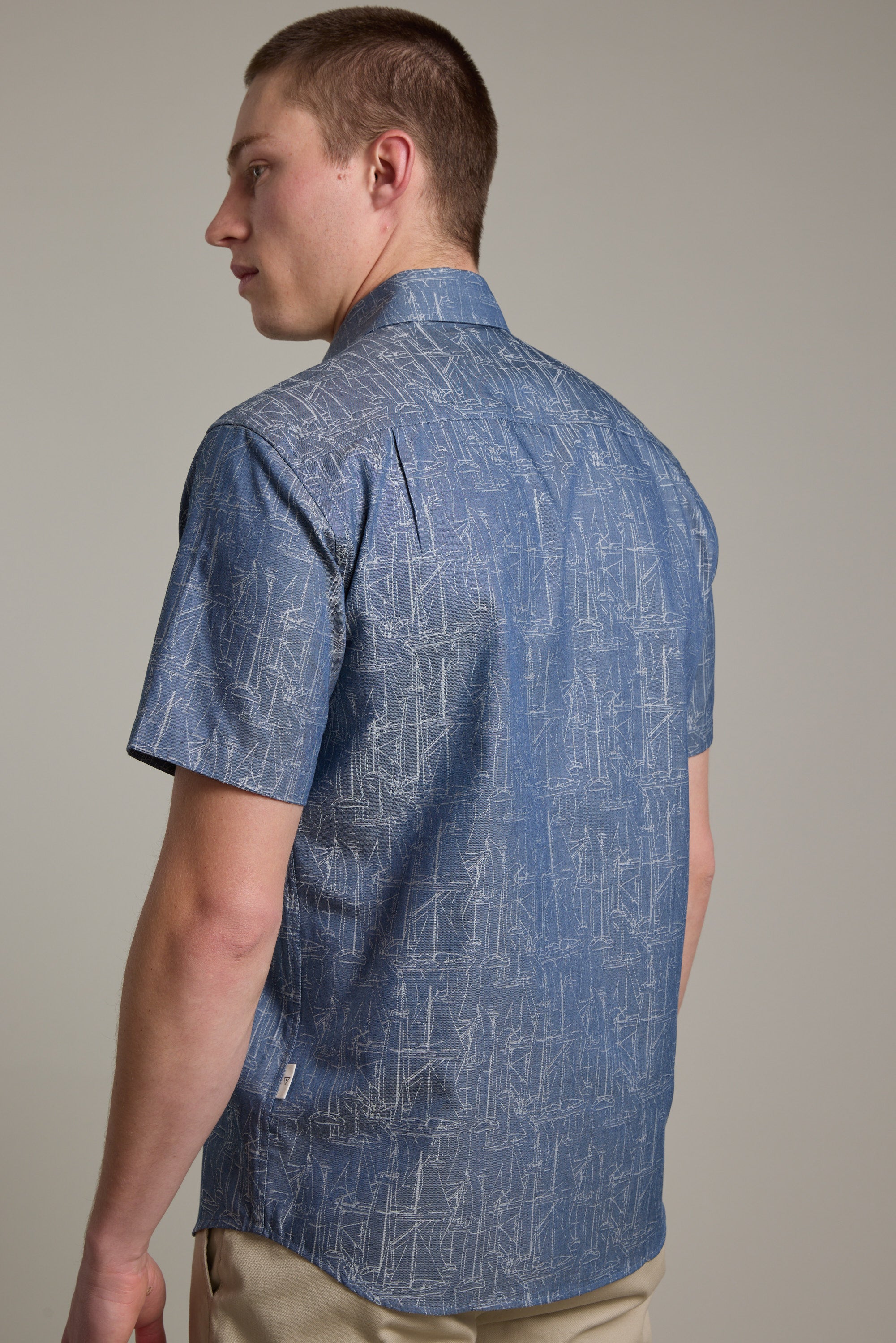 A person wearing the Barkers Yacht Jacquard Shirt, featuring short sleeves and a blue yacht print, with beige pants, stands facing away from the camera and looks over their left shoulder against a plain background.