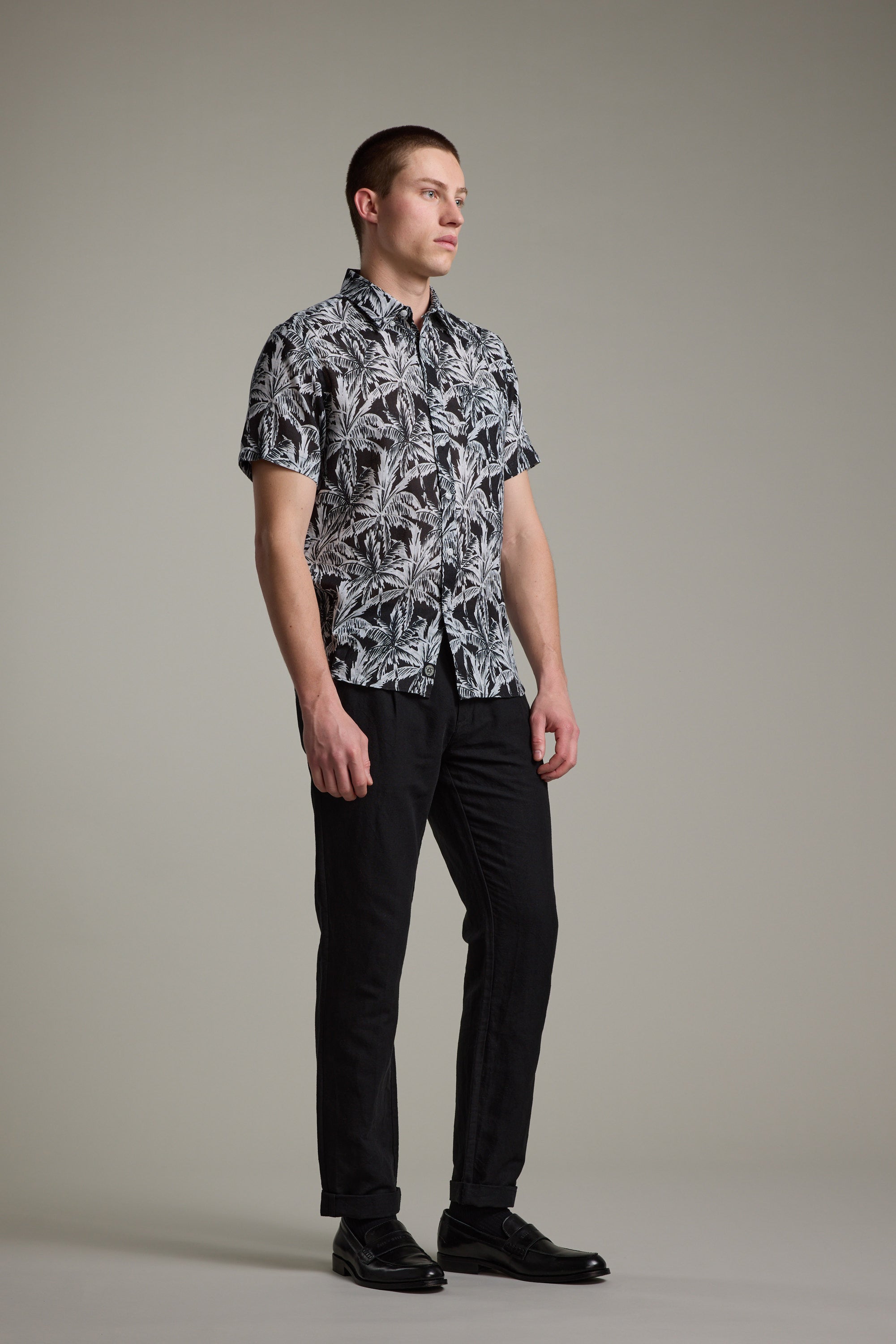 A man stands against a plain background, wearing the Barkers Madison Print Shirt—a black and white Hawaiian style in a cotton–linen blend—paired with black pants and dress shoes, looking slightly to the side.