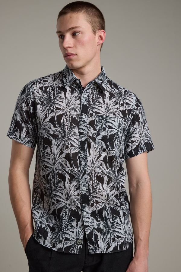 A young man with short hair models the Barkers Madison Print Shirt—a black-and-white palm leaf design in a linen-cotton blend. He stands against a plain background, one hand in his pocket and gazing to the side.