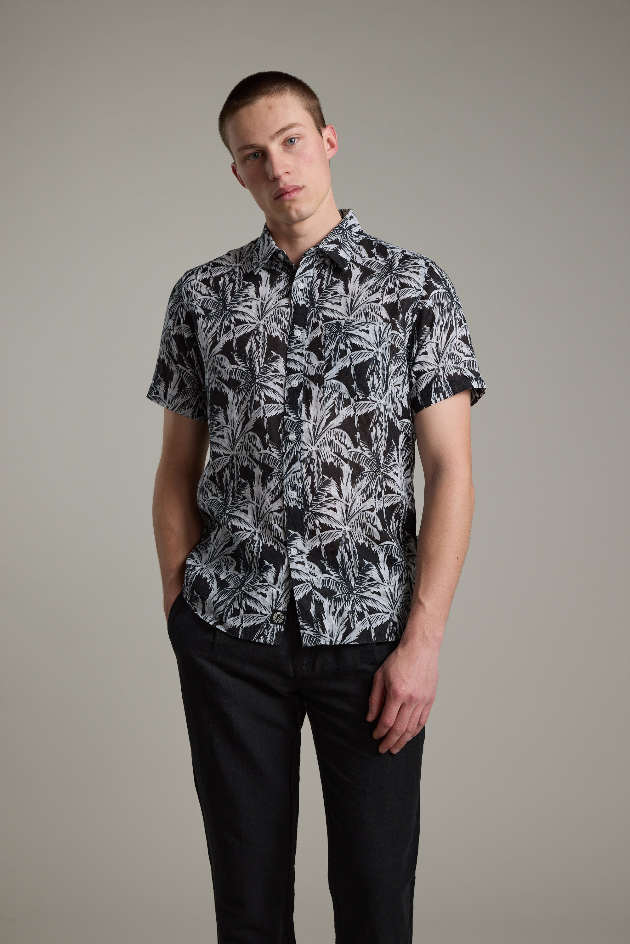 A young man stands against a plain background, wearing the Barkers Madison Print Shirt—a short-sleeved, black cotton–linen blend with a white tropical leaf pattern—and black pants. He has short hair, a relaxed expression, and one hand in his pocket.